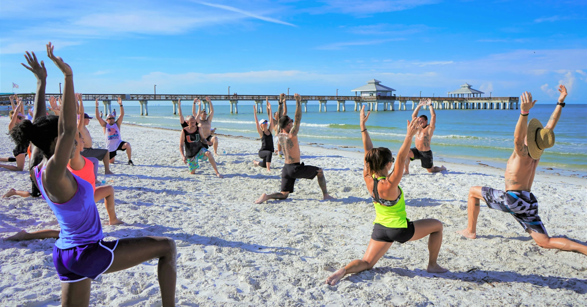 Group of people doing a yoga session on a sandy beach with a wooden pier and ocean in the background.