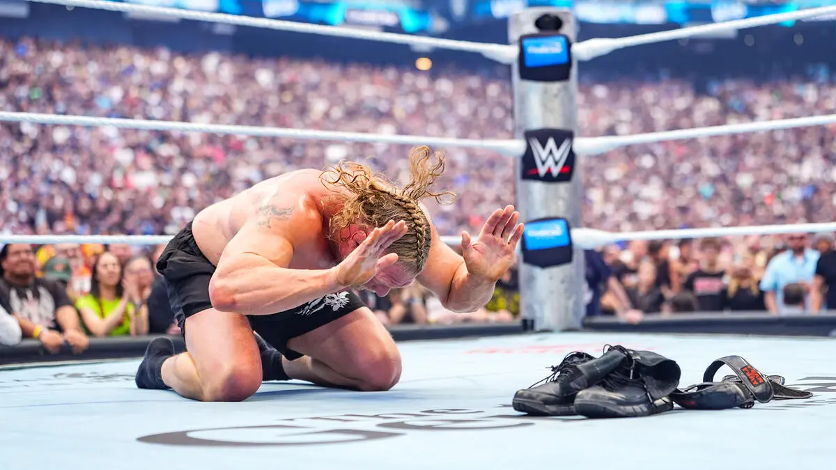 WWE wrestler kneels on the ring mat in pain, arms up to shield his head, crowd blurred in the background; boots and belt lie nearby on the mat.