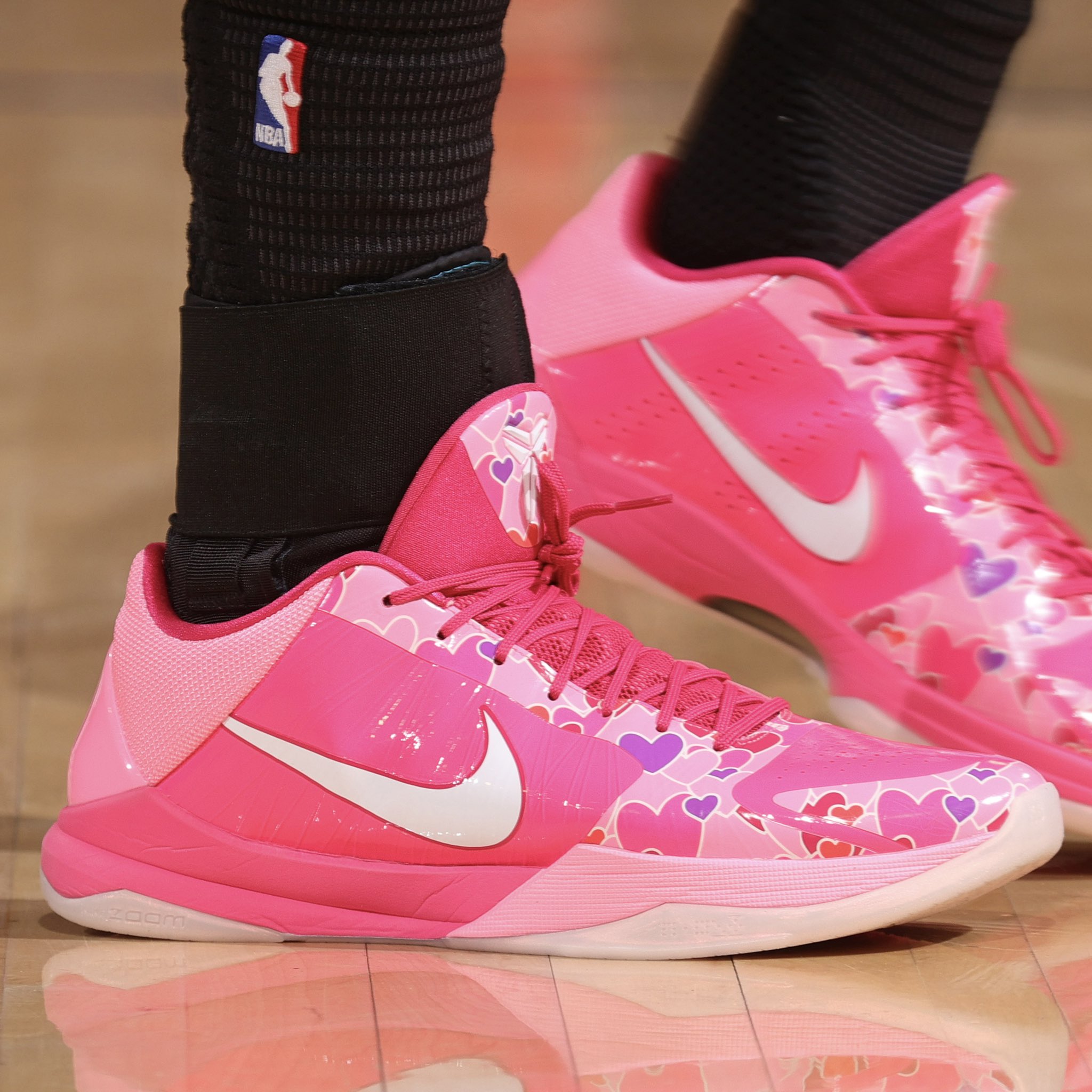 Bright pink Nike basketball shoes with white swoosh and heart-pattern detail, worn with black socks on a wooden court.