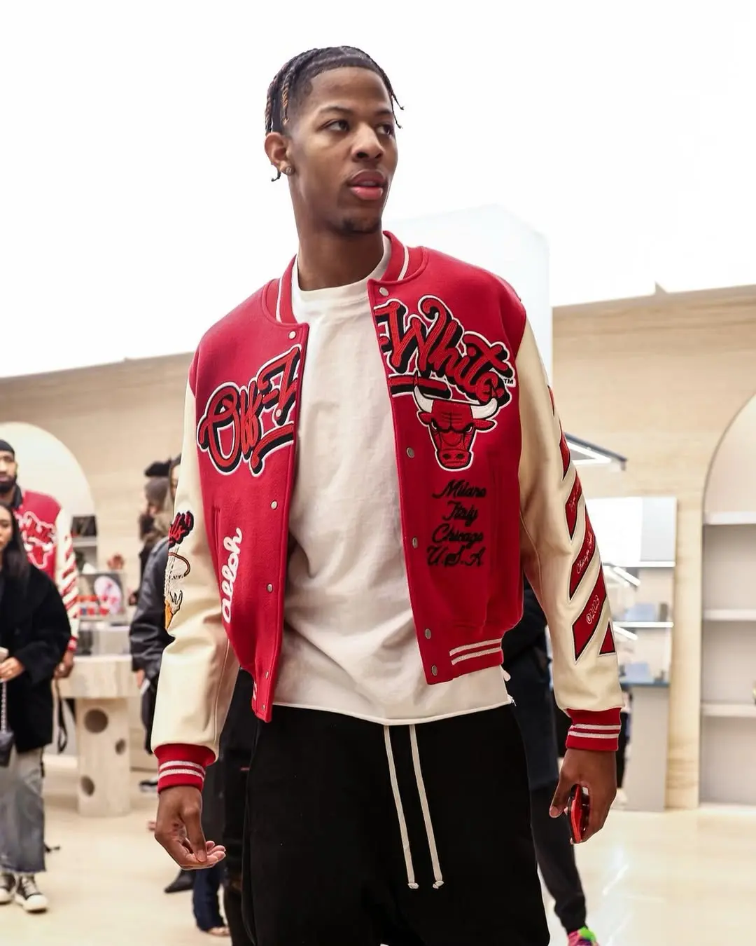Young man wearing a red and cream varsity jacket with bold graphics, white T‑shirt, and black drawstring pants in a shopping mall.