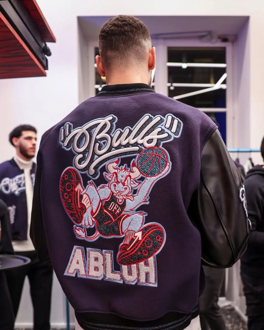 Person seen from behind wearing a purple varsity jacket with a large embroidered cartoon rabbit playing basketball on the back, indoors among others.