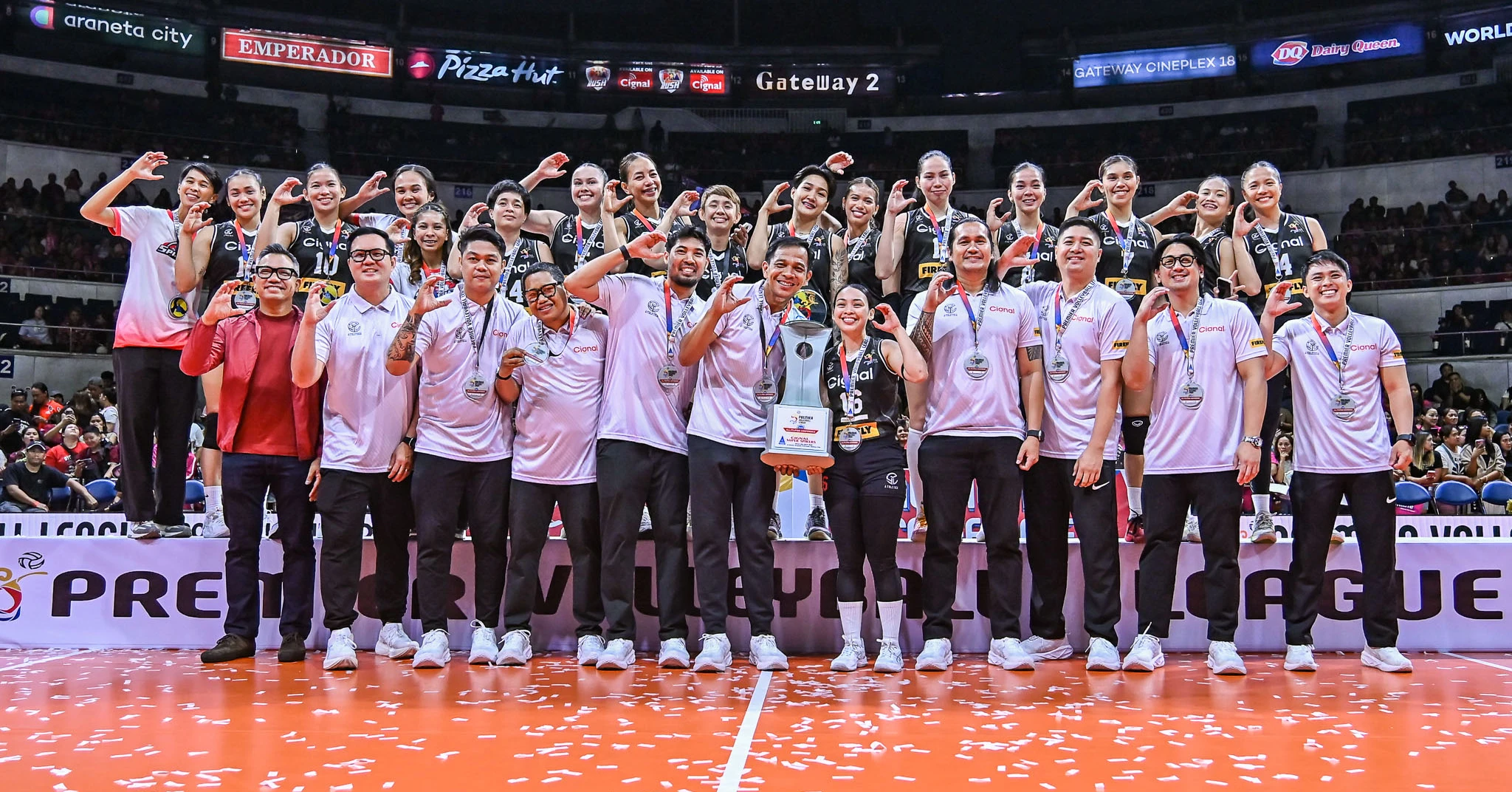 Group of athletes posing on a court with medals and a trophy after a win, confetti on the floor and an arena crowd in the background