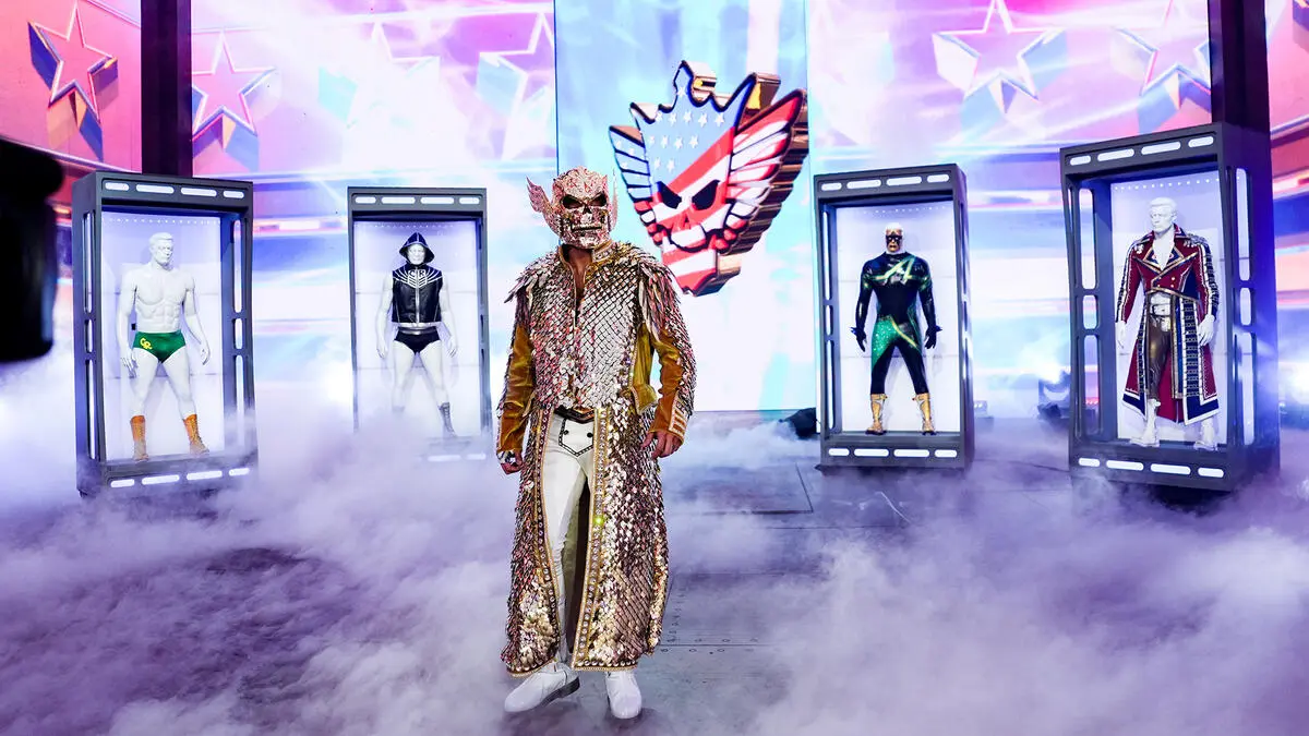 Cody 2 - the game Masked performer in a gold sequined robe walks through fog toward the audience, with five hero costumes on display behind.