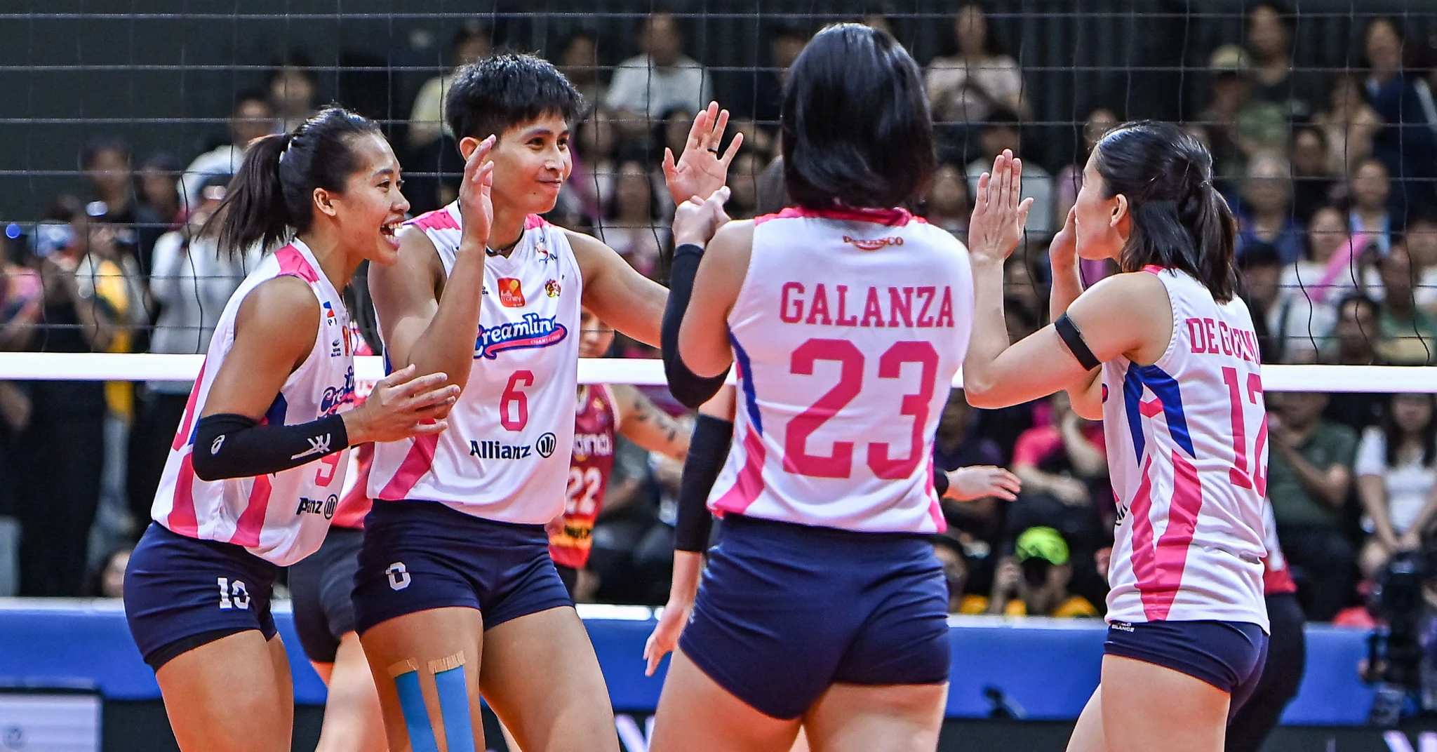 A group of female volleyball players in pink-and-white uniforms high-fiving each other on the court, net in the background, crowded stands behind them.