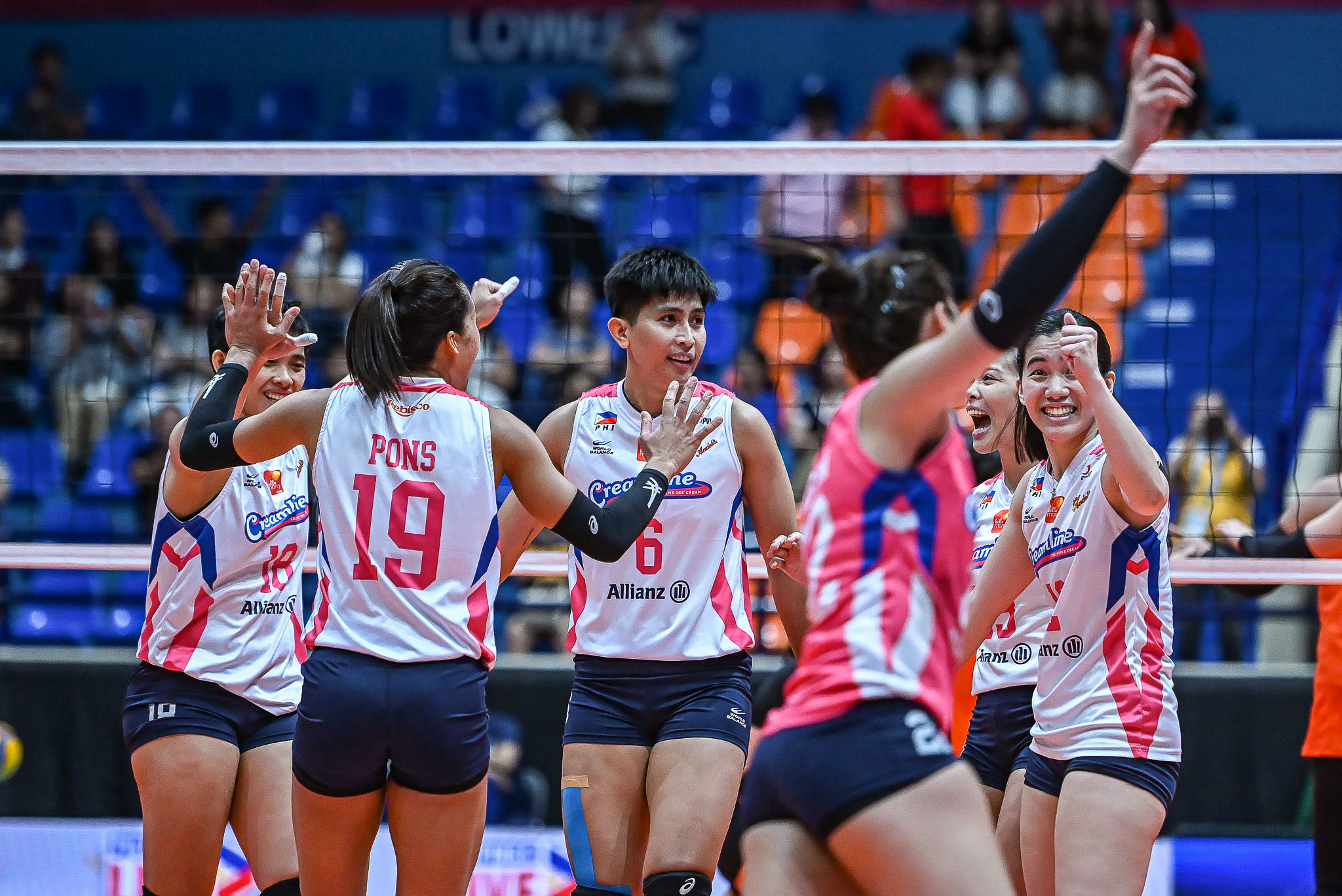 Female volleyball players in white and pink jerseys celebrate a point with high-fives near the net on a crowded indoor court.
