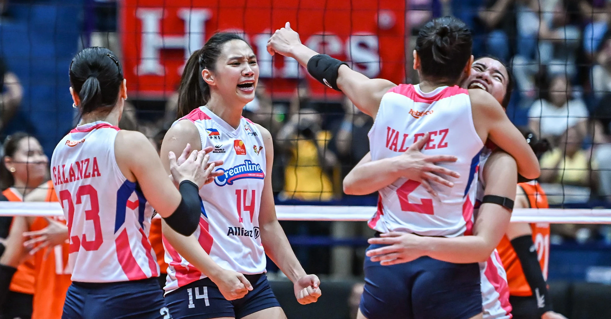 Volleyball players in white-pink uniforms celebrate a point, hugging near the net.