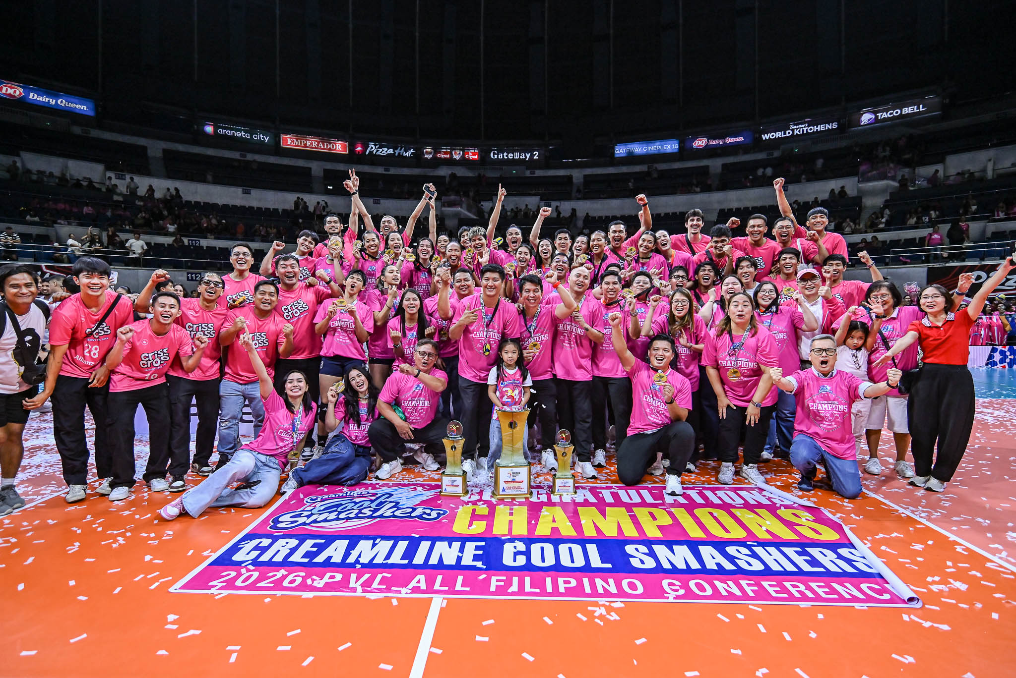 Creamline - the game Large group of people in pink shirts pose with trophies on an orange indoor court, confetti falling as they celebrate a championship victory.