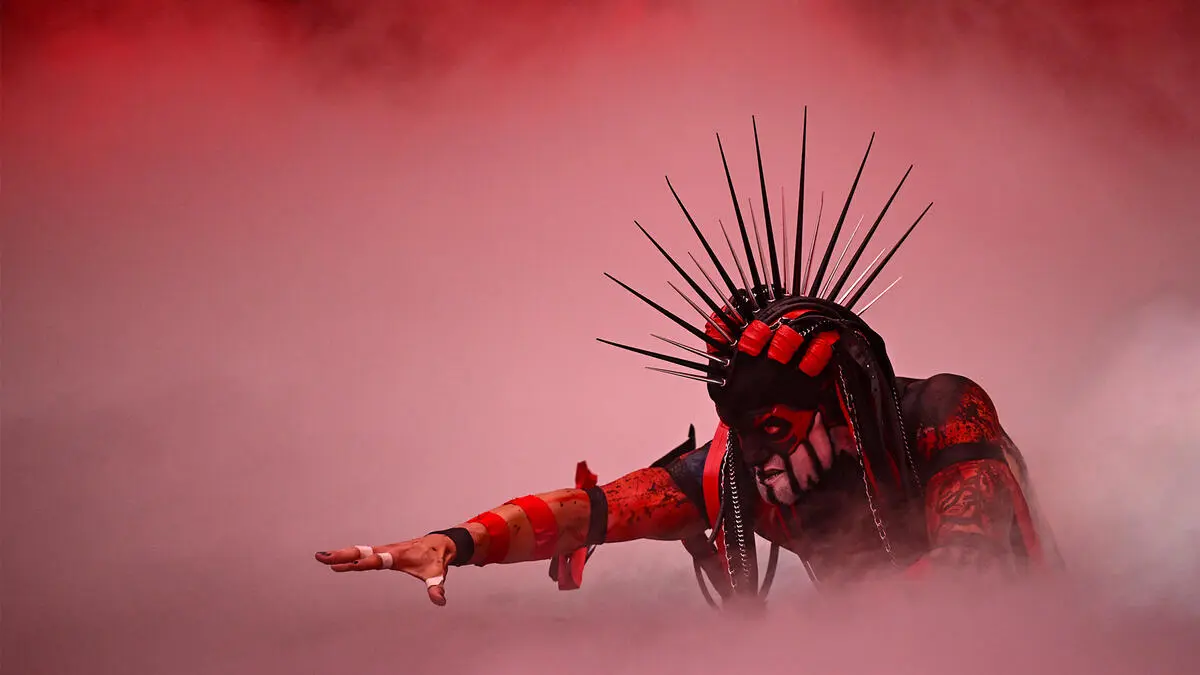 Person in black and red makeup with a tall spiked headdress reaches forward through pink mist at a stage performance.