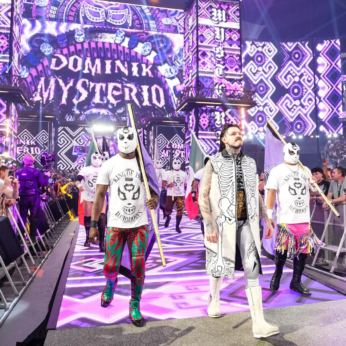 Dom - the game Masked wrestlers with staffs march down a purple-lit entrance ramp toward the ring. Fans line the barrier in the foreground, vivid arena graphics behind them.