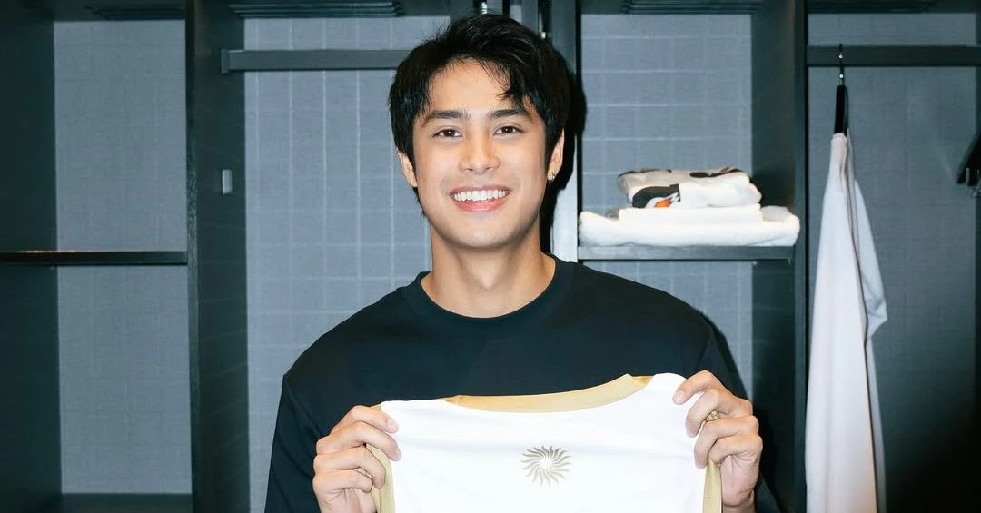 Smiling man in a black shirt holds up a white T‑shirt with a gold sun emblem in a locker room.