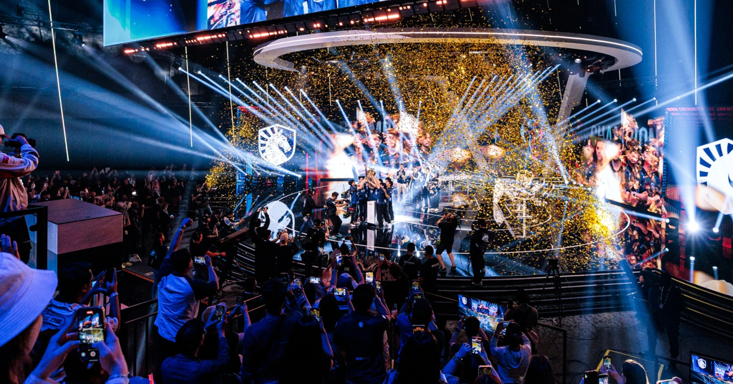 Esports team celebrates on a bright stage with confetti as fans film and cheer from the audience.