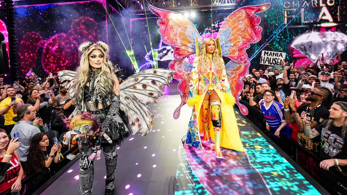 Flair n bliss - the game Two performers with large colorful butterfly wings walk a runway as a cheering crowd watches from both sides.