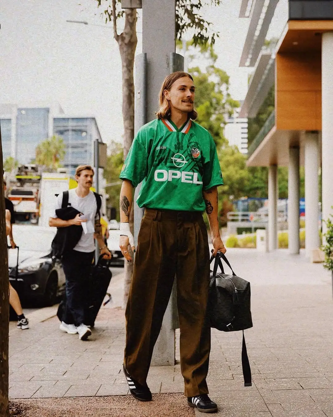 Irvine - the game Man with shoulder-length hair in a green Opel soccer jersey and brown pants stands on a city sidewalk, holding a black bag.