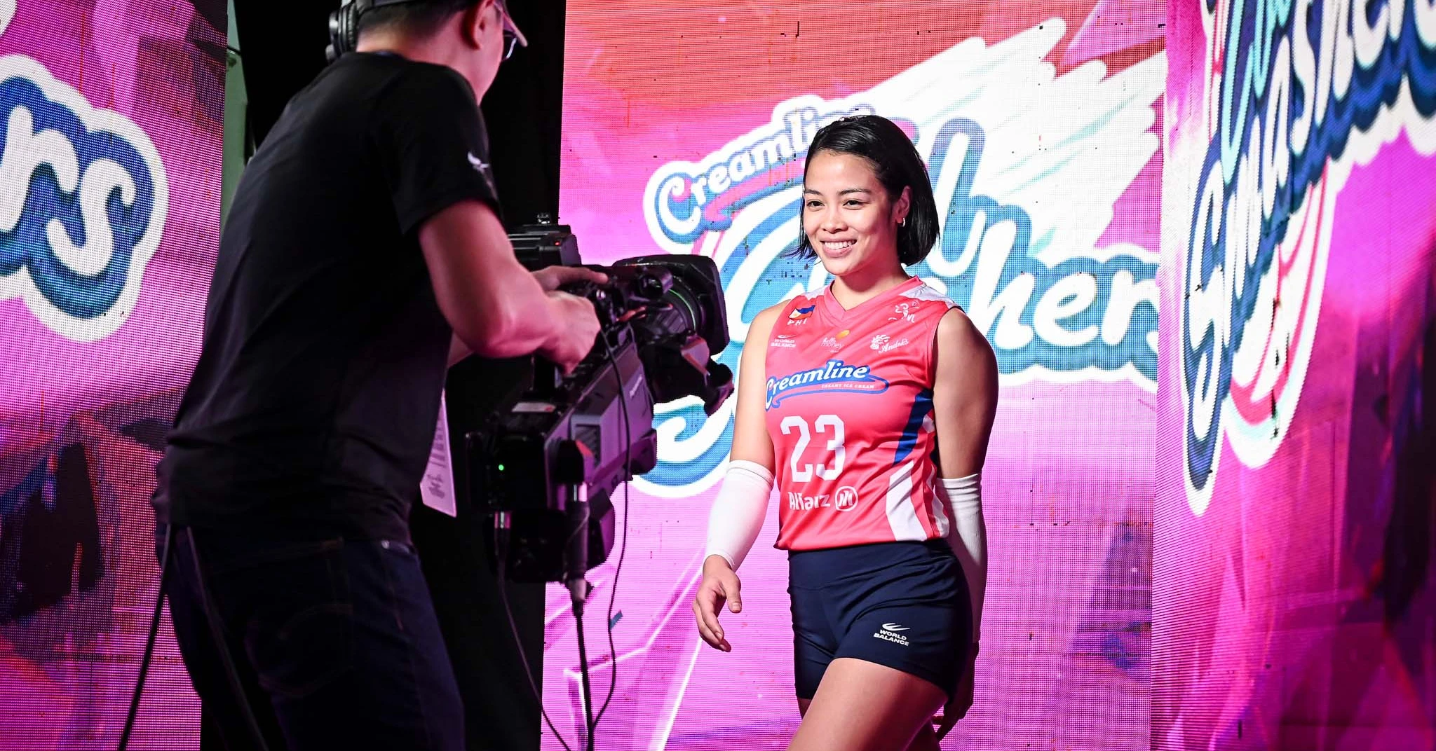 Female volleyball player in a pink jersey (#23) smiles as a cameraman films her on a colorful stage backdrop.