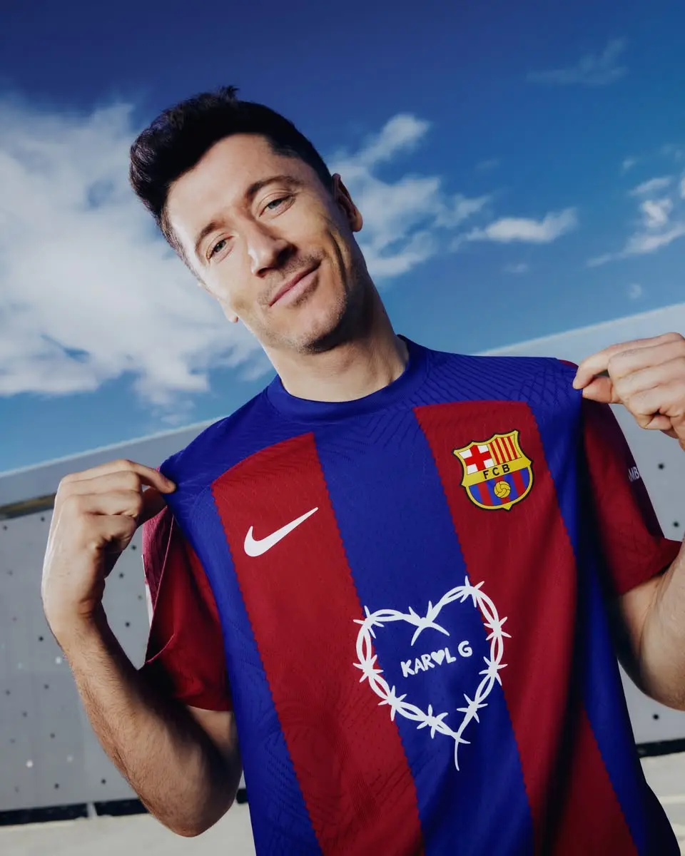 Smiling man wearing a FC Barcelona jersey outdoors, lifting the shirt to show the crest against a blue sky.