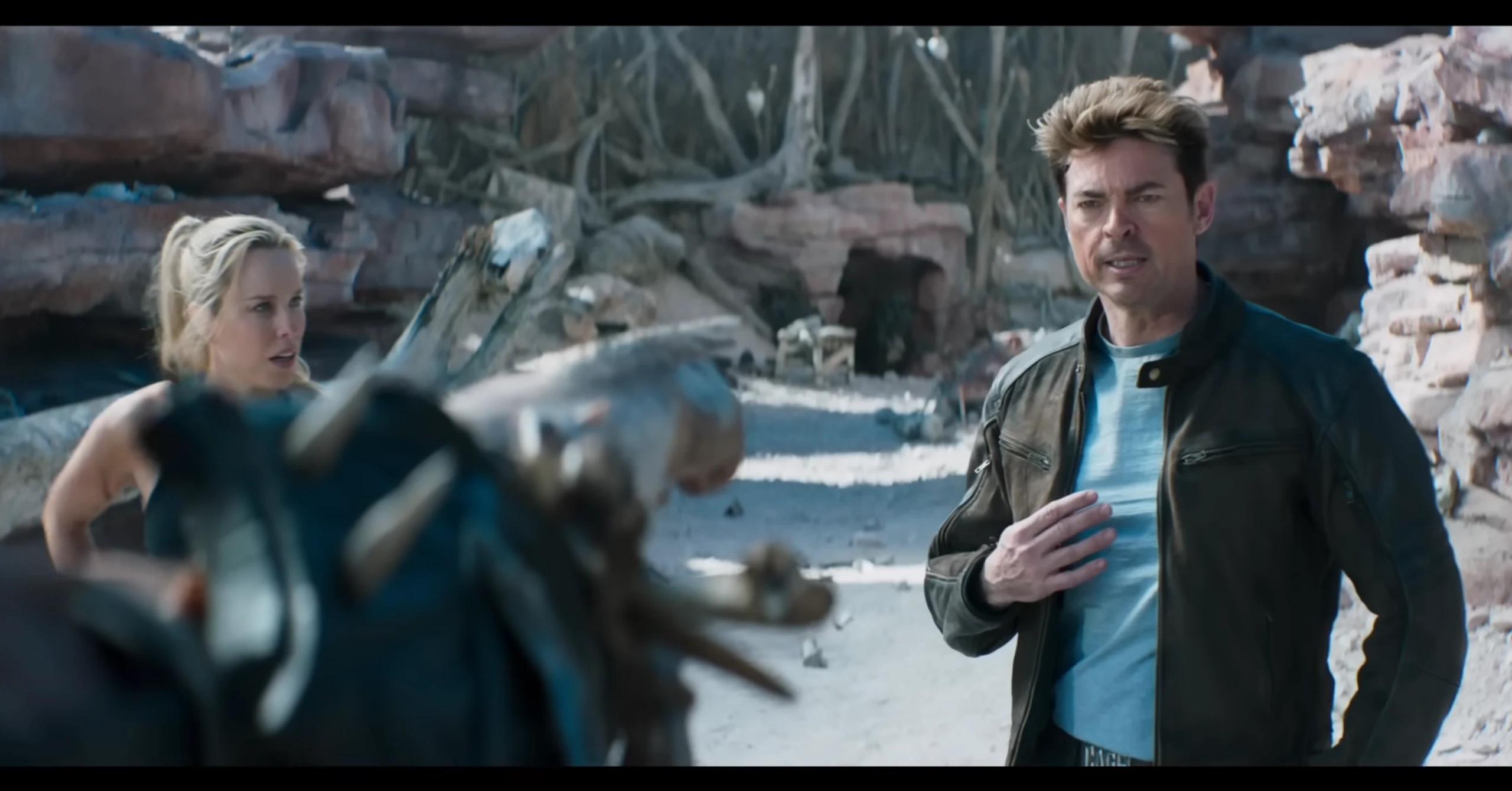 Man in a brown jacket on a rocky beach, speaking to a blonde woman off to the left, with a blurred foreground figure in the foreground.