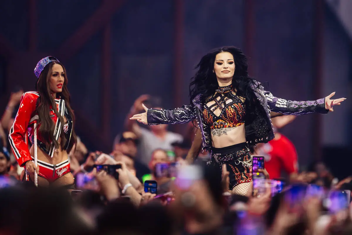 Two female performers on a crowded stage, one in a black-and-flame crop top and studded jacket with arms outstretched, the other in a red-and-white outfit, entertaining the audience.