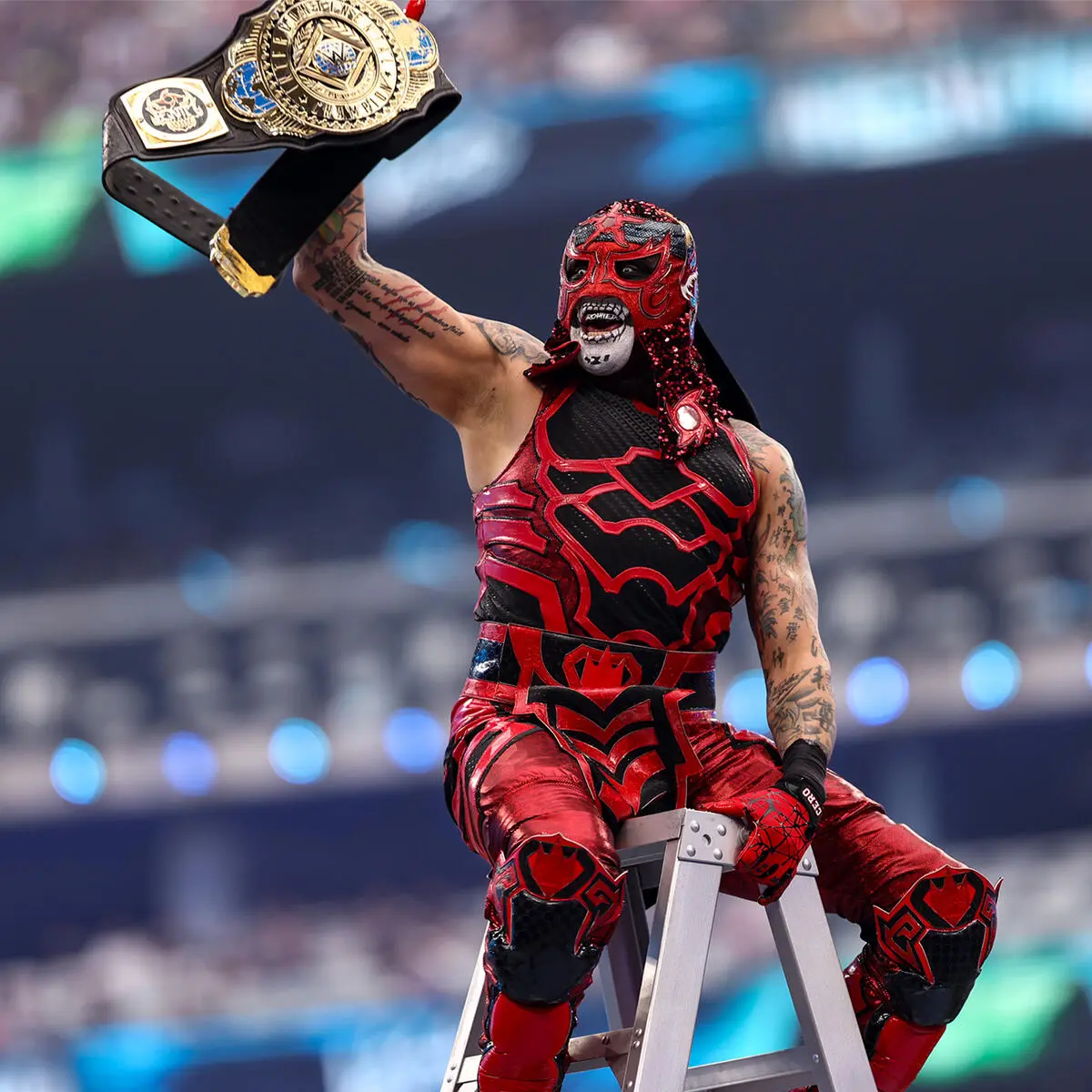 Masked professional wrestler in red and black gear raises a championship belt while perched on a ladder in the ring stadium