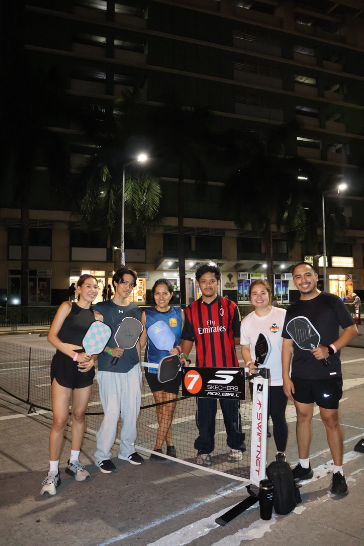 Pickleball-ortigas - the game Six people pose with pickleball paddles on a nighttime outdoor court, smiling in front of a net beneath streetlights.