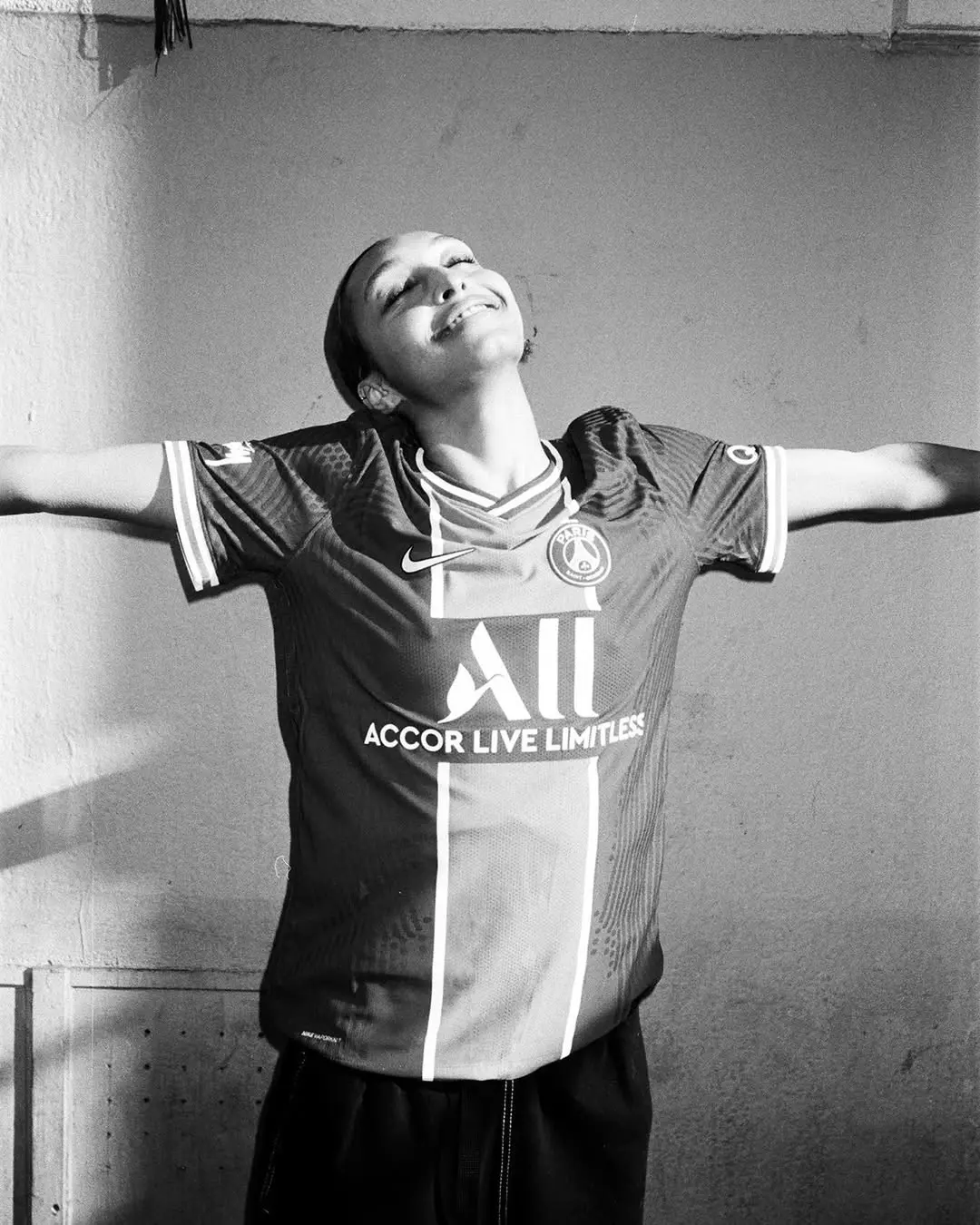 Person in a PSG football jersey stands with arms outstretched and head tilted back, smiling against a plain wall.