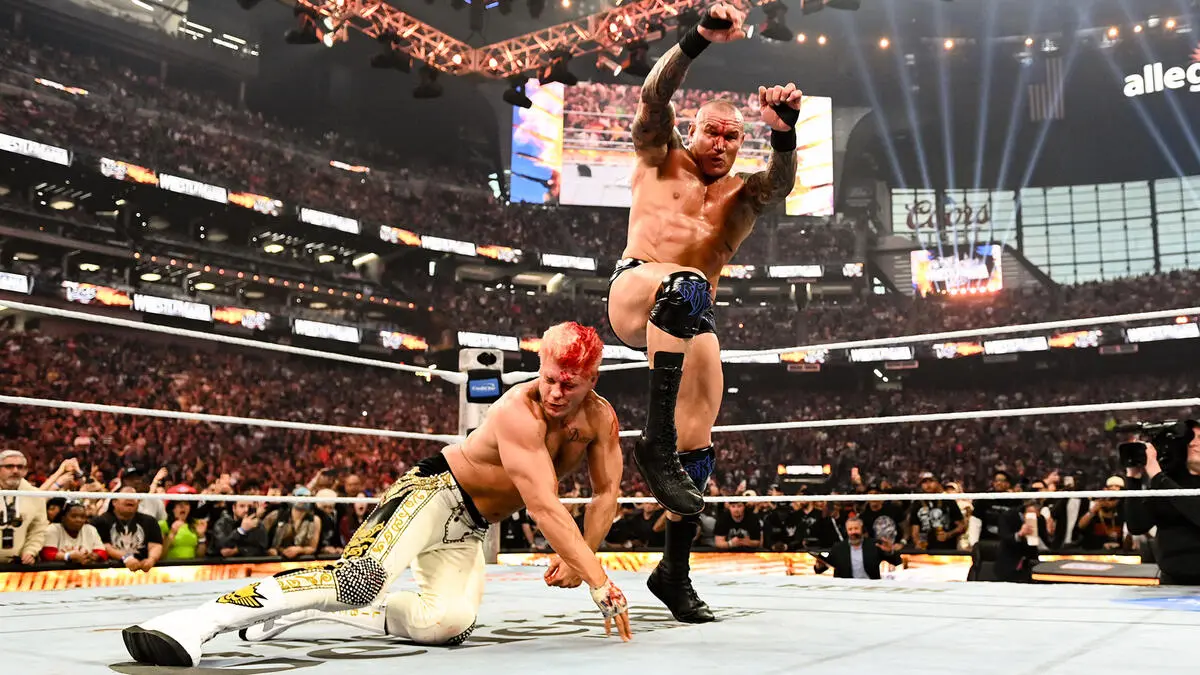 Two wrestlers battle in a crowded arena; one leaps into the air toward an opponent on the mat as the audience cheers.