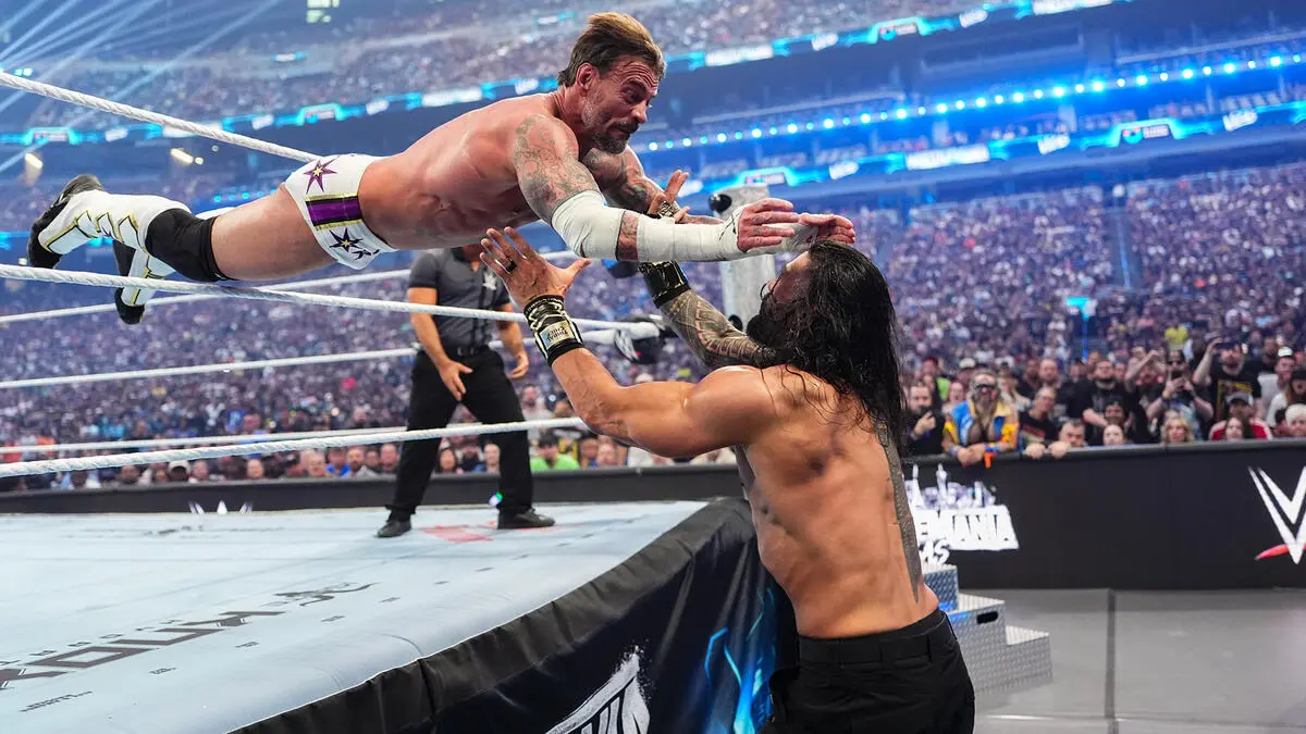 Wrestler in white trunks jumps into a high-flying move toward an opponent reaching up from the ring edge as a large crowd watches.