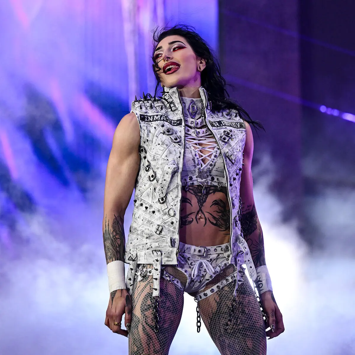 Rhea ripley - the game Female rock performer in a white studded vest and fishnet tights on stage, singing under purple lights.