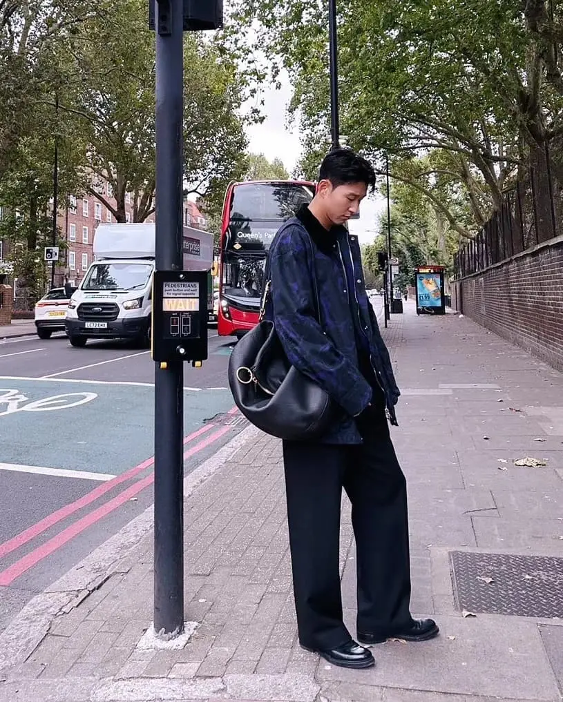 Son - the game Young man in a dark blue jacket and black pants stands on a city sidewalk, carrying a black shoulder bag, with traffic and a red bus behind him.