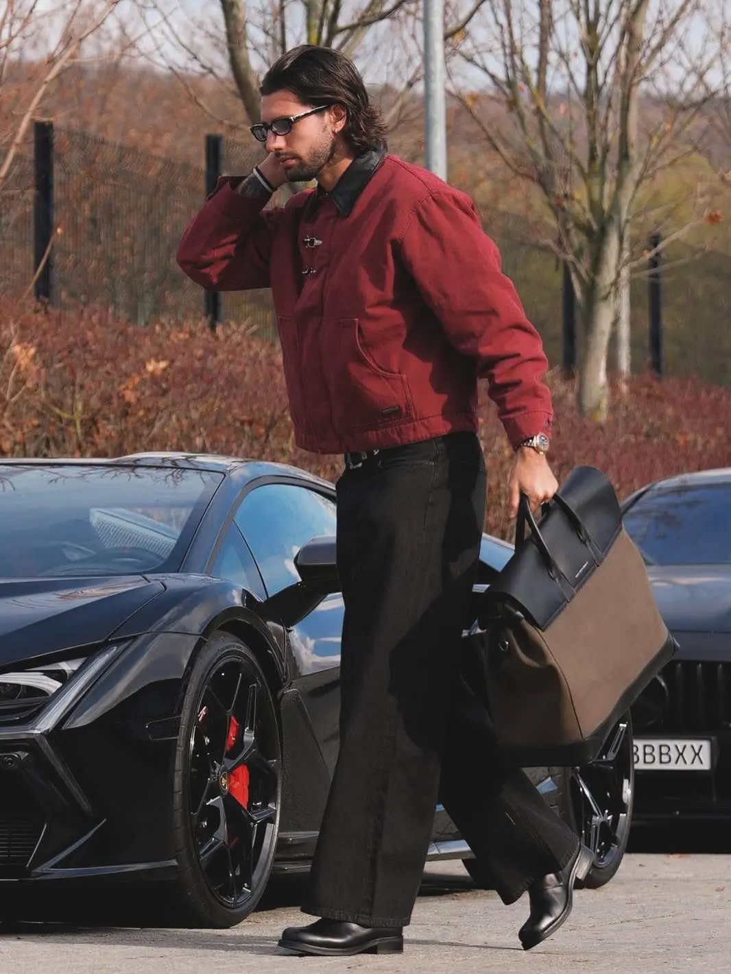 Szobo - the game Man in a burgundy jacket and sunglasses carries a large brown-and-black bag beside a black sports car in a parking lot.