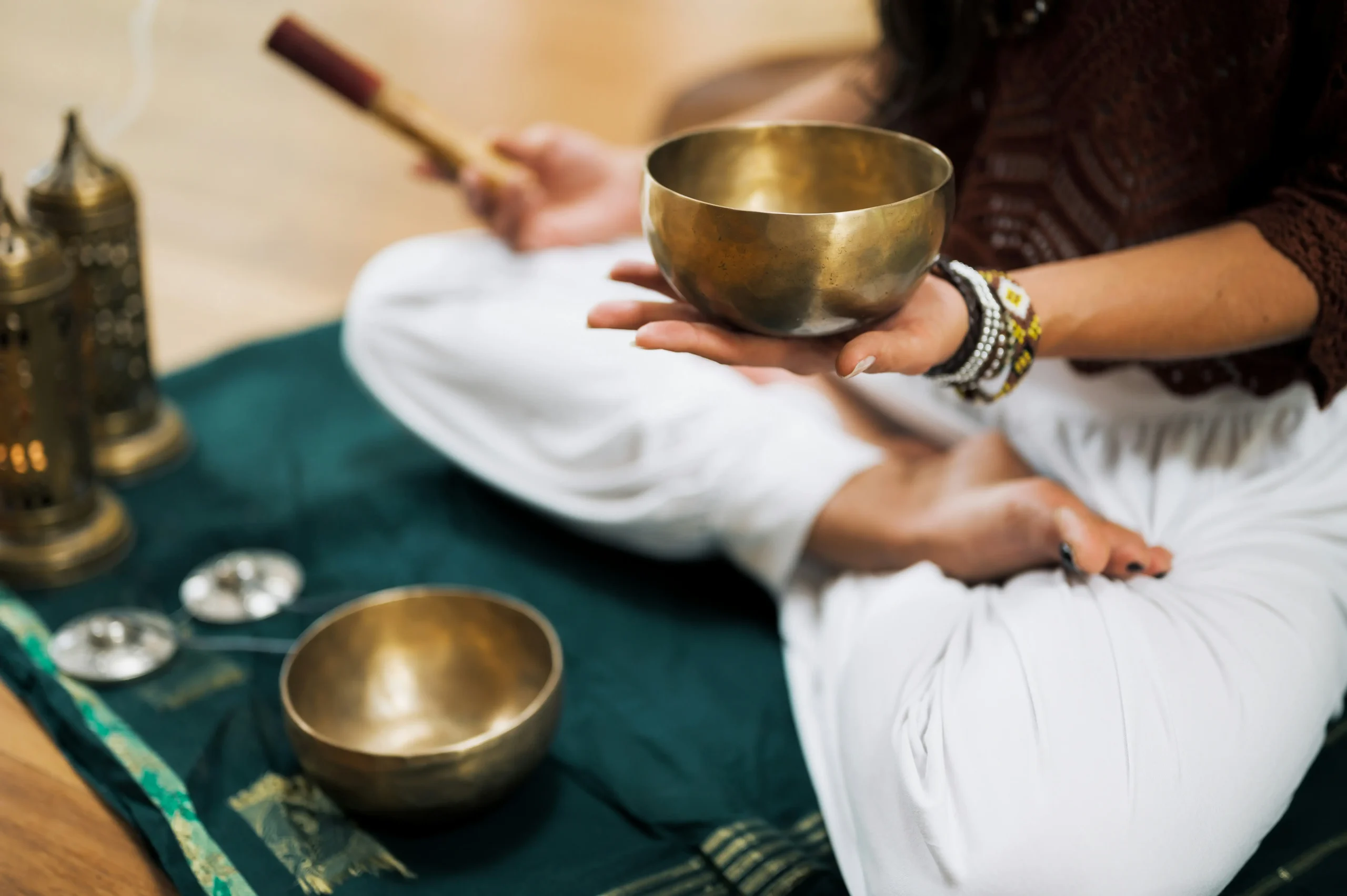 Thlt-lcx-hn4wyhoaeic-unsplash - the game Person wearing bracelets holds a brass singing bowl during a sound therapy setup, with other bowls on a teal cloth nearby.