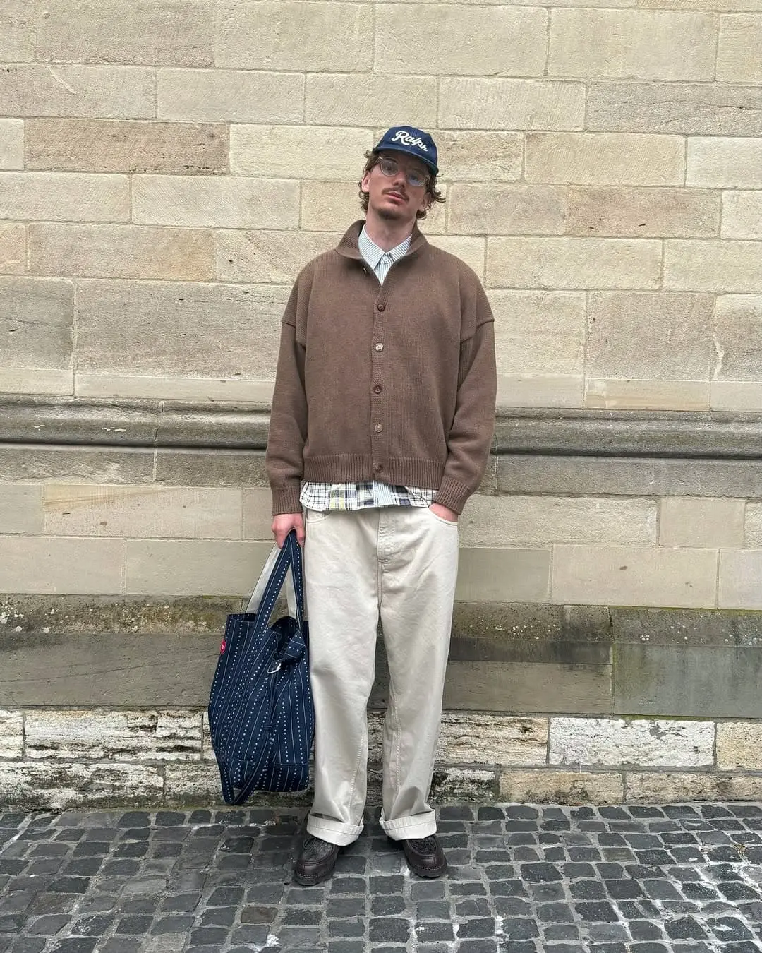 Woltemade - the game Man in a brown cardigan and plaid shirt stands against a stone wall, holding a navy striped tote bag.