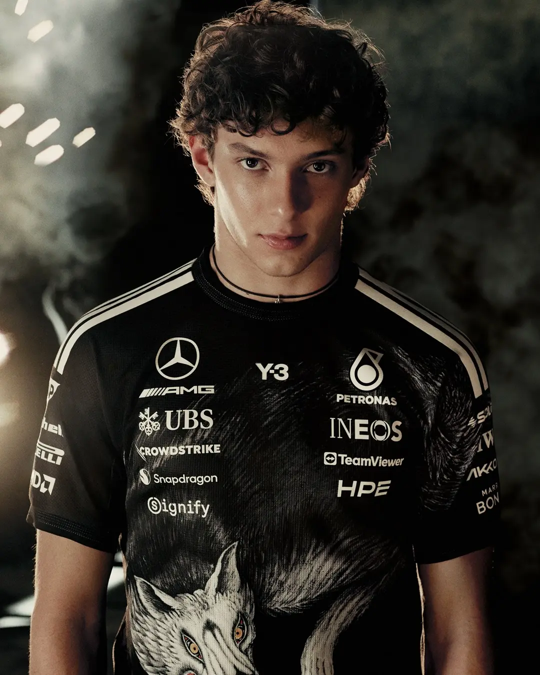 Young man in a black Formula 1 team jersey with sponsor logos (Mercedes, Petronas, INEOS) and a wolf graphic on the shirt, against a dark background.