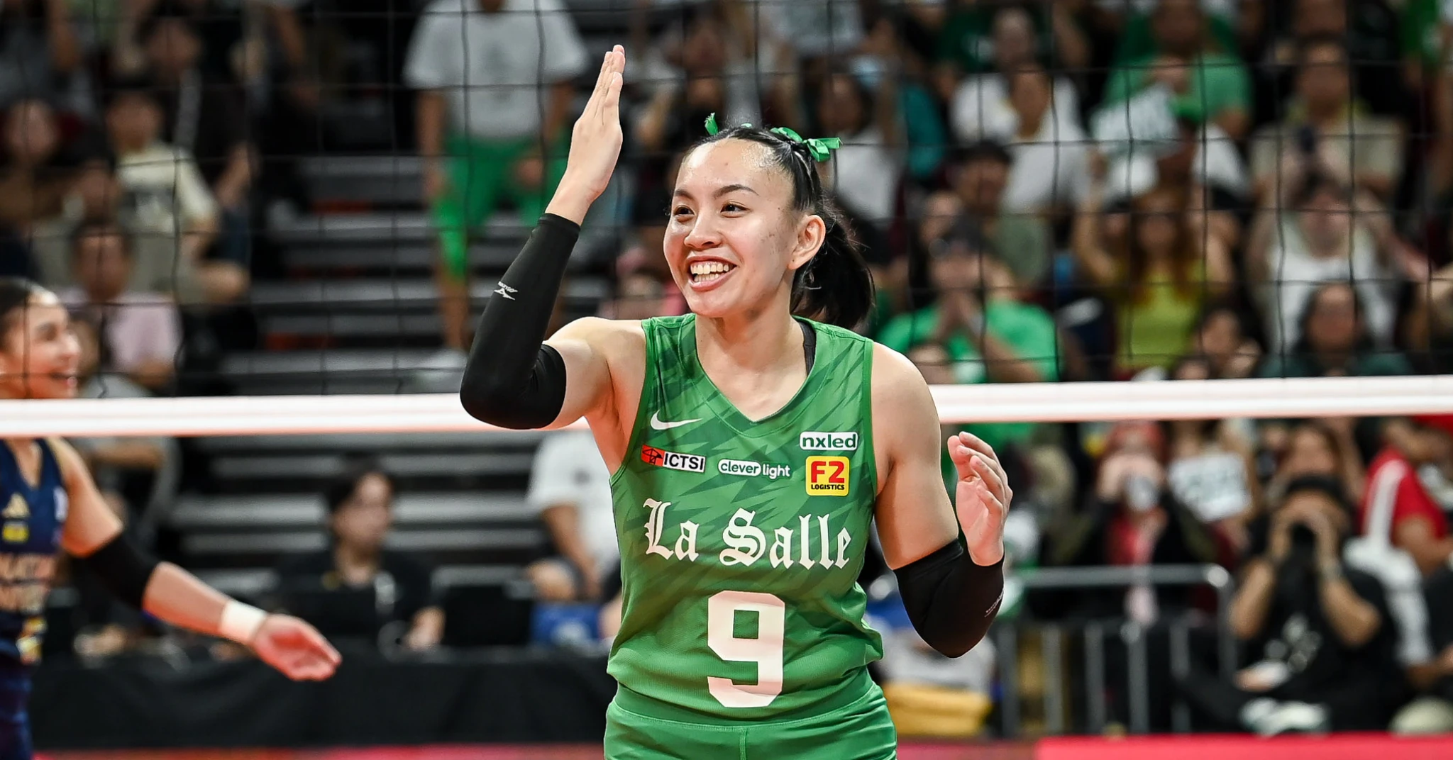 Female volleyball player in a green La Salle jersey raises her hand in celebration on the court; net is visible in the foreground with a cheering crowd behind.