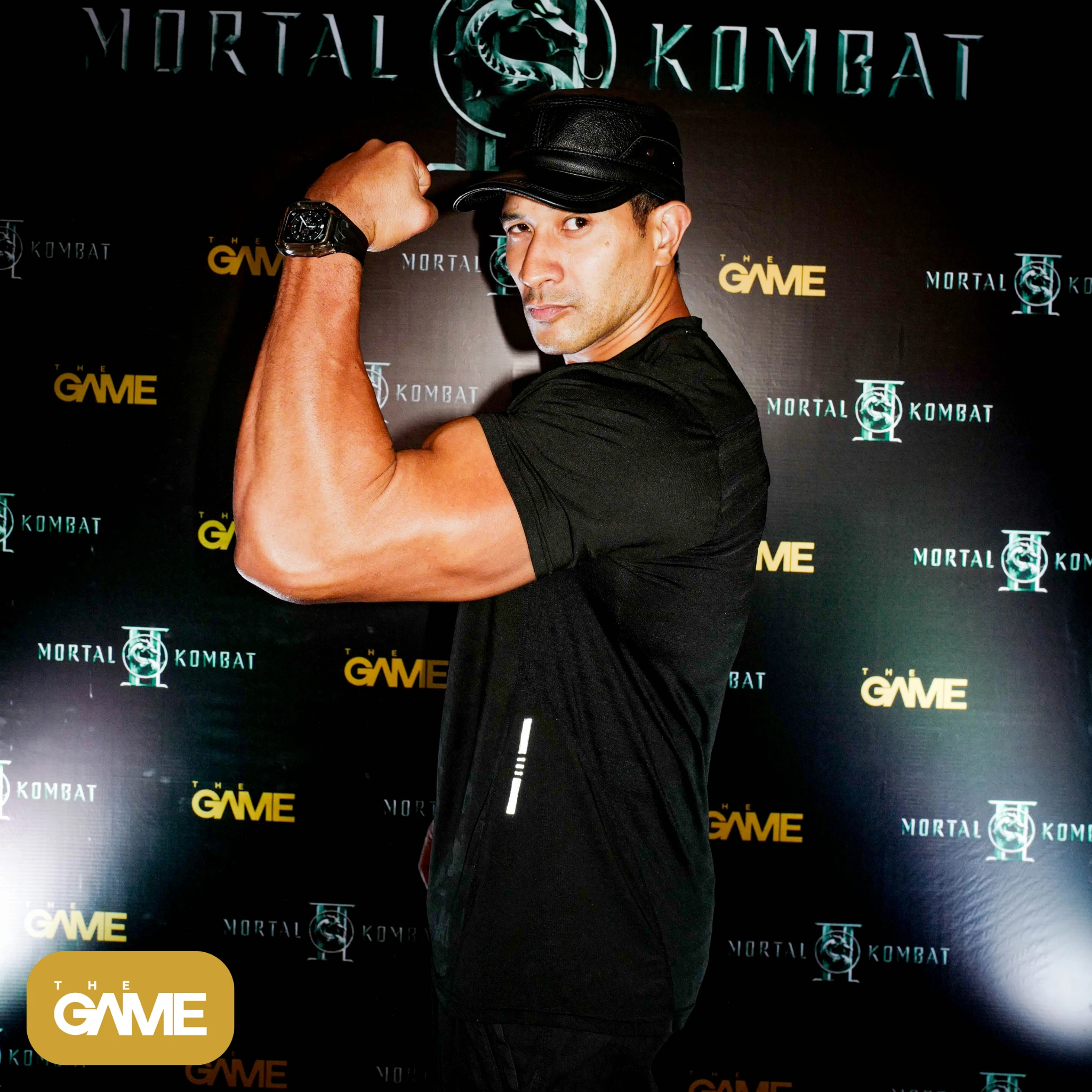 4 6 - the game Muscular man in a black t-shirt and cap flexes his bicep, posed in front of a Mortal Kombat backdrop with logos.