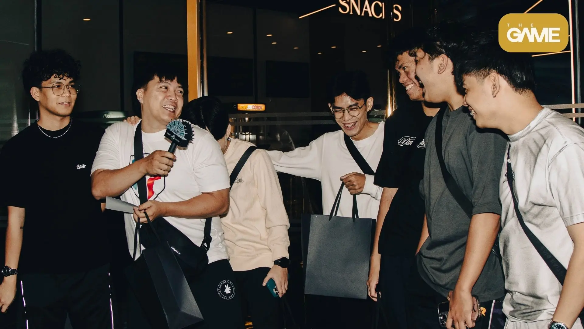 5 - the game Group of friends smiling and chatting indoors, a man holds a microphone and a paper as others gather around him, casual wear and bags visible, logo in corner.
