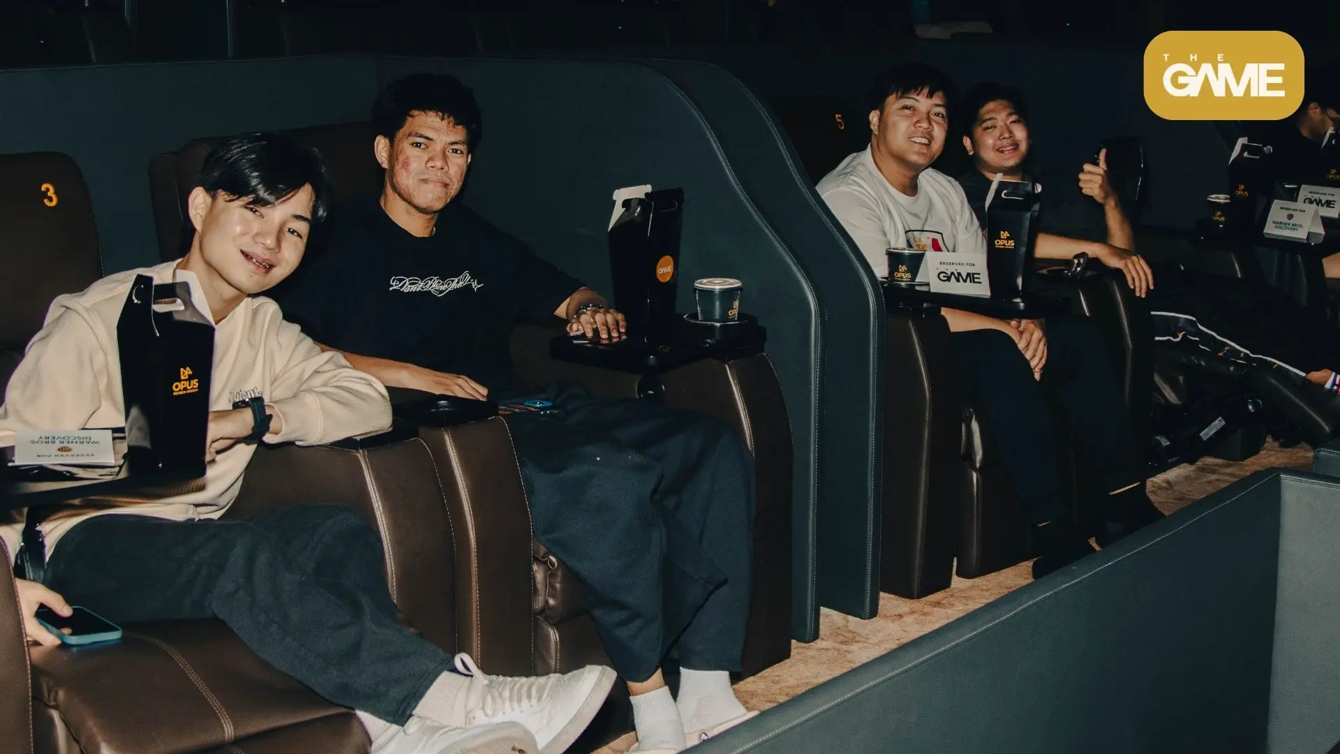 Ap bren - the game Group of four people seated in dark, plush theater seats at a gaming event, with drinks on small tables and reserved signs nearby; a yellow THE GAME logo appears in the top-right corner.