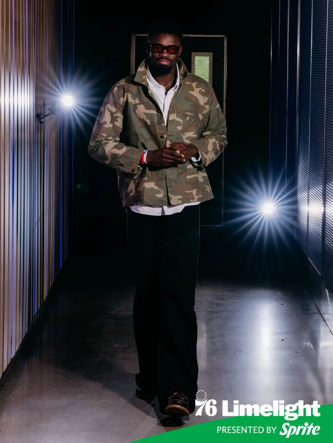 Adem bona - the game Man in a camouflage jacket and sunglasses stands in a dim, spotlight-lit hallway, posing for a Limelight event photo.