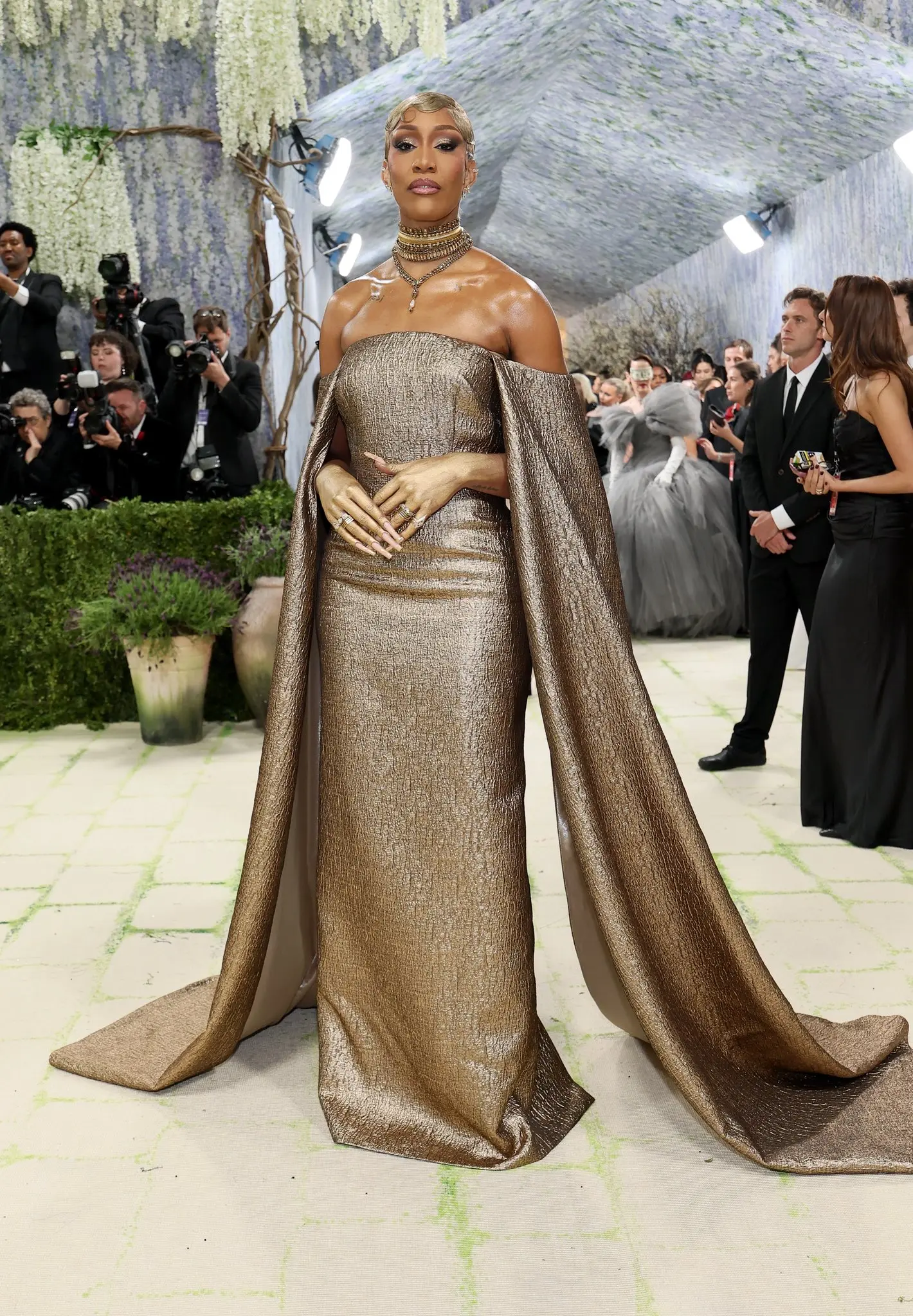 Aja wilson - the game Glamorous woman in a metallic gold off-shoulder gown with a long cape, on a red carpet event surrounded by photographers.
