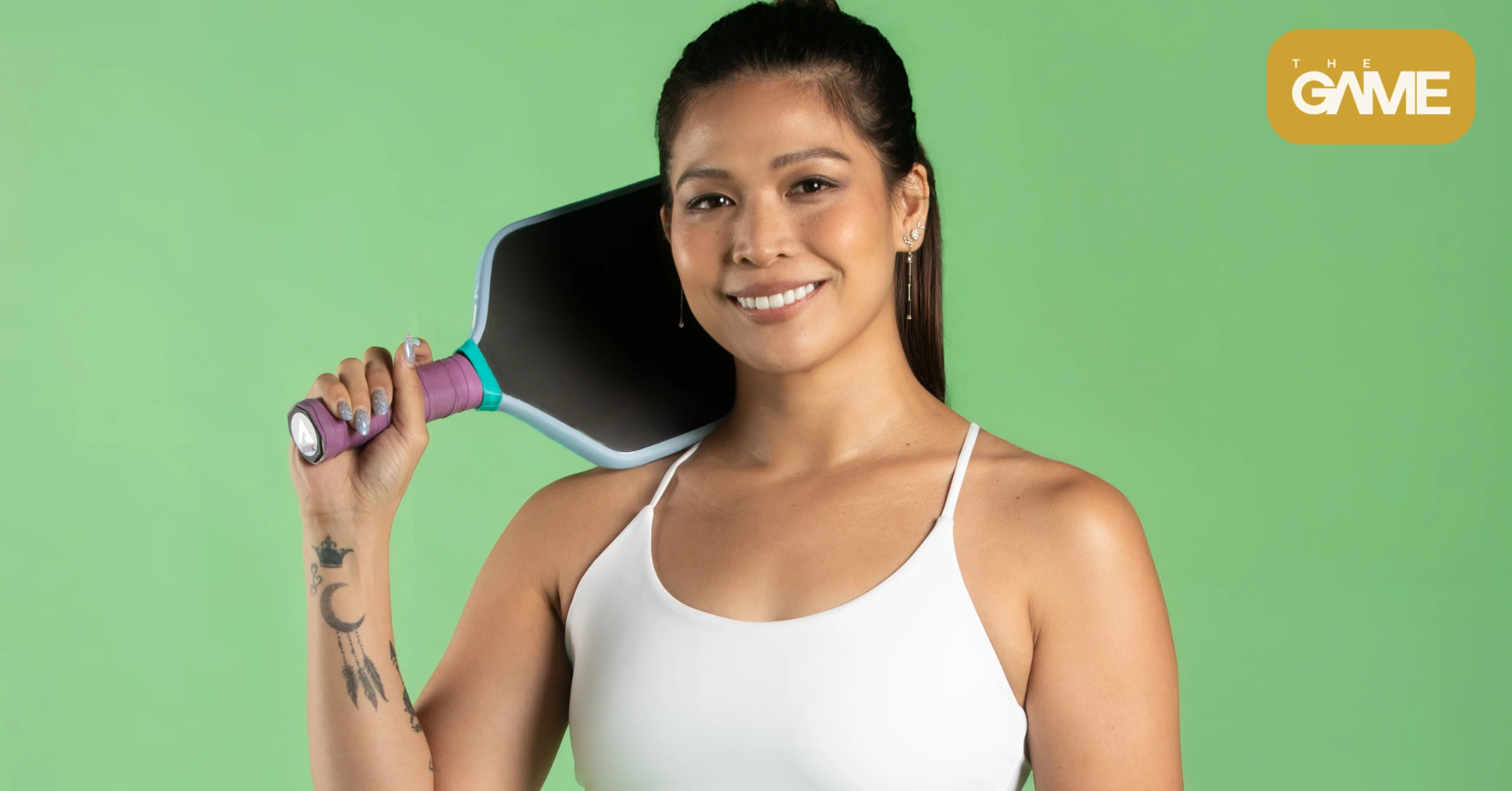 Smiling woman in a white top holds a pink paddle racket over her shoulder against a green background; The Game logo in the top-right corner.