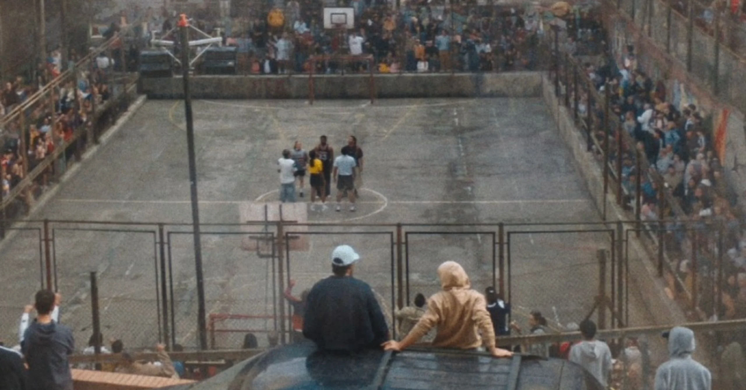 People gathered on an outdoor basketball court surrounded by a large crowd behind a chain-link fence.