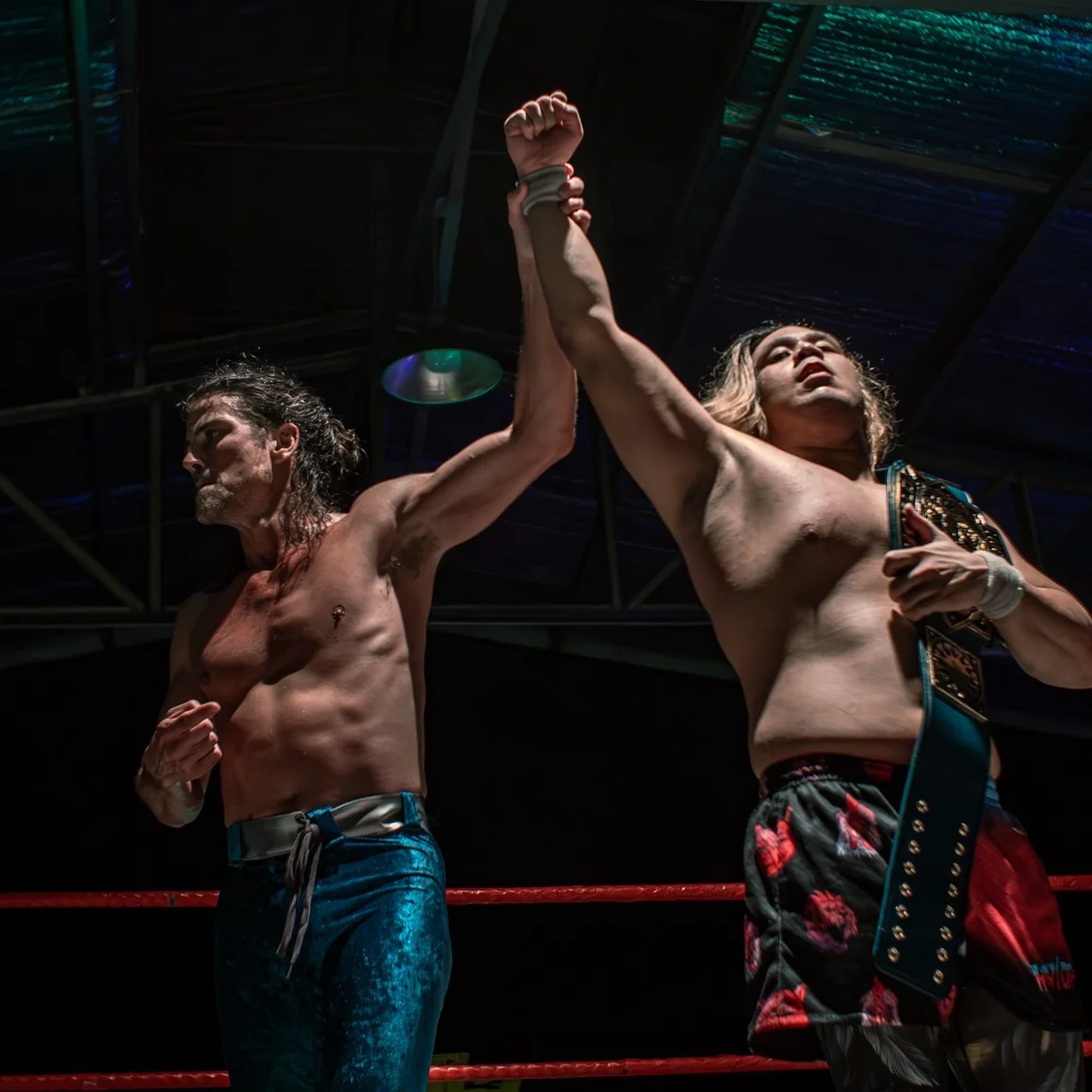 Brian kendrick - the game Two shirtless wrestlers raise their arms in victory inside a boxing ring; one wears a championship belt over his shoulder, the other clenches a fist, both under dramatic lighting.