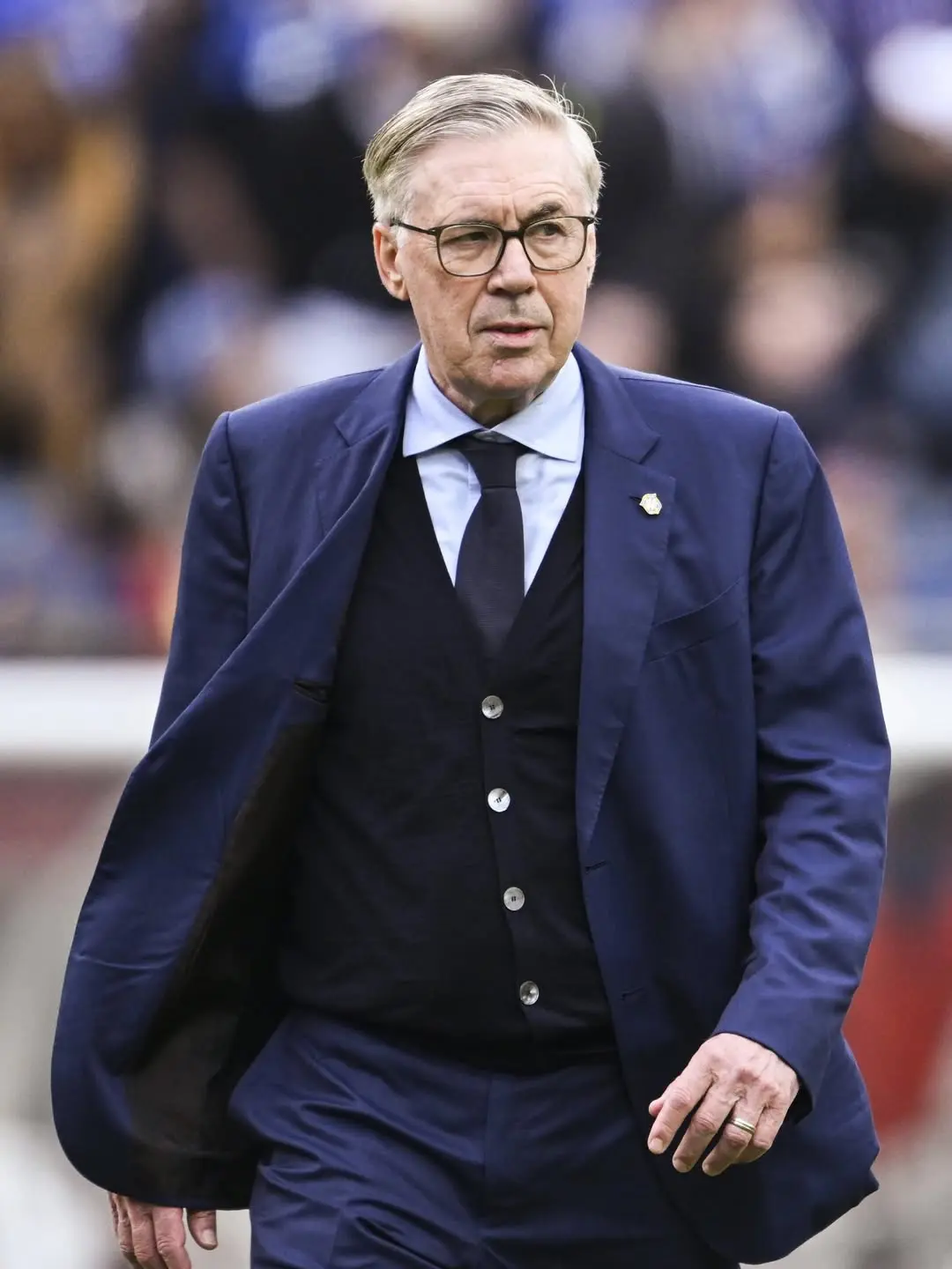 Carlo ancelotti - the game Man in a navy suit and vest, wearing glasses, walking on a sports field with a blurred crowd in the background.