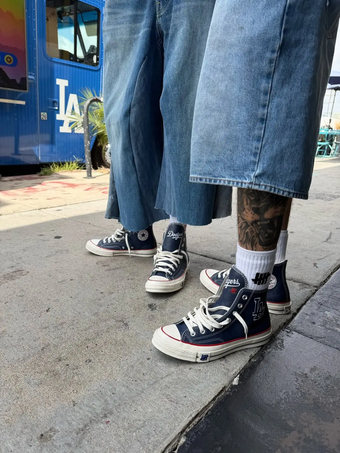 Three people in long denim skirts and navy high-top sneakers stand on a sidewalk, one with a tattooed leg visible on the right side.