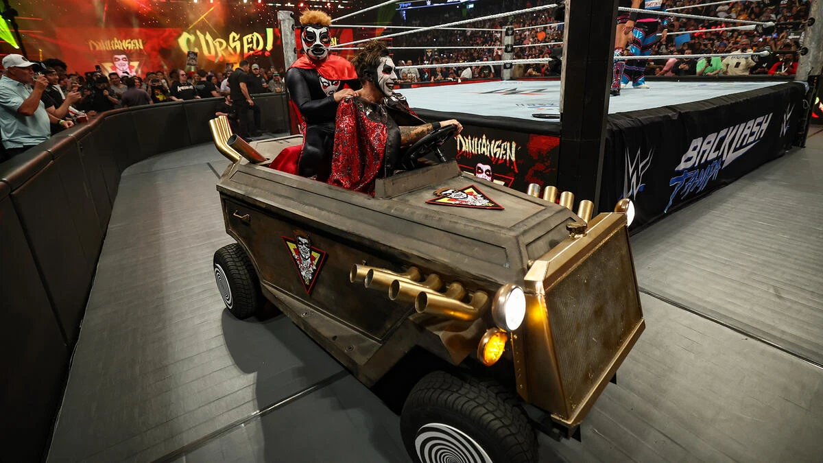 Danhausen - the game Two costumed wrestlers with clown makeup ride in a rugged, armored car near the wrestling ring, with fans in the background.”,