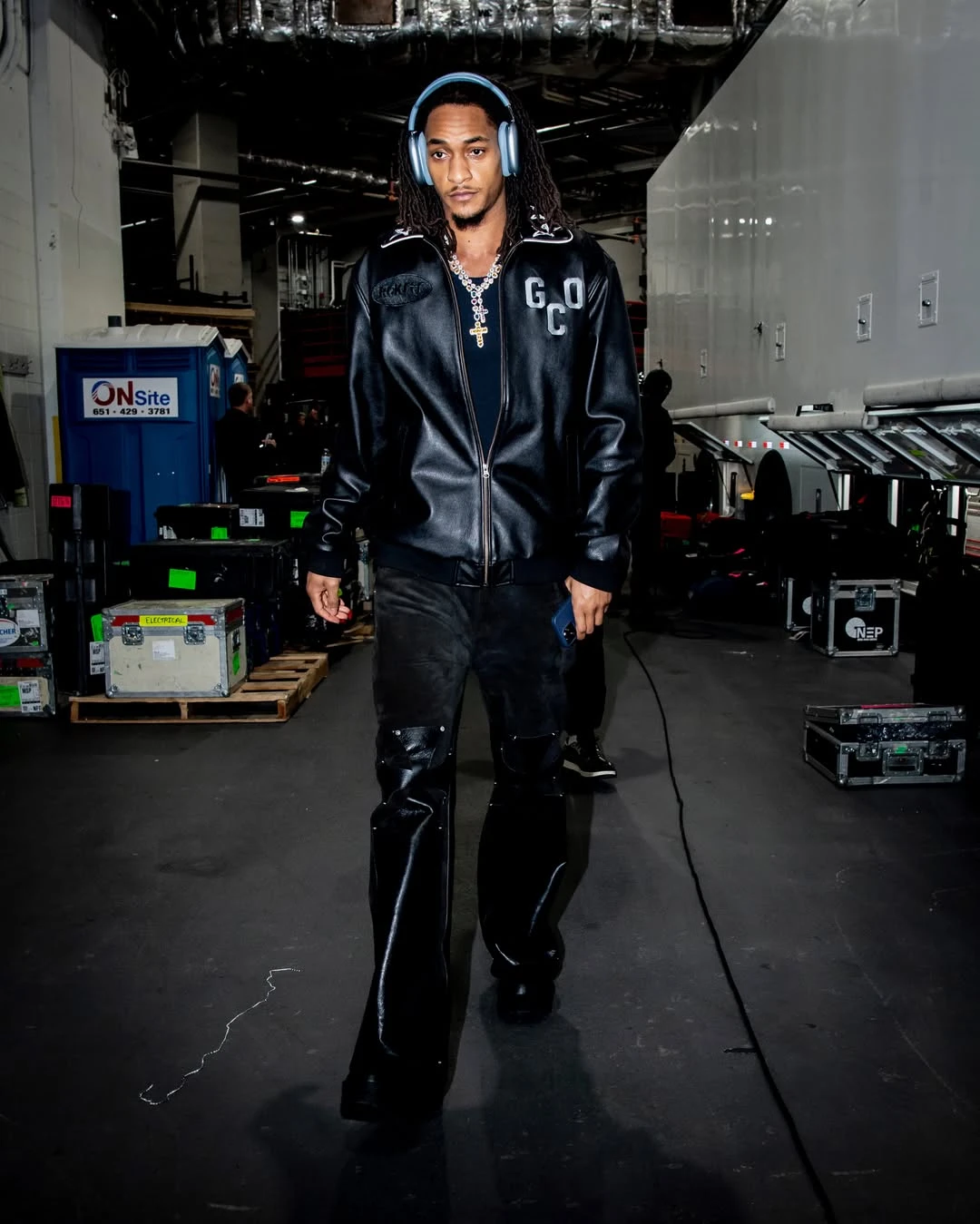 Devin vassell - the game Man walking backstage in a black leather jacket and blue headphones, surrounded by equipment cases and cables in a tech venue.