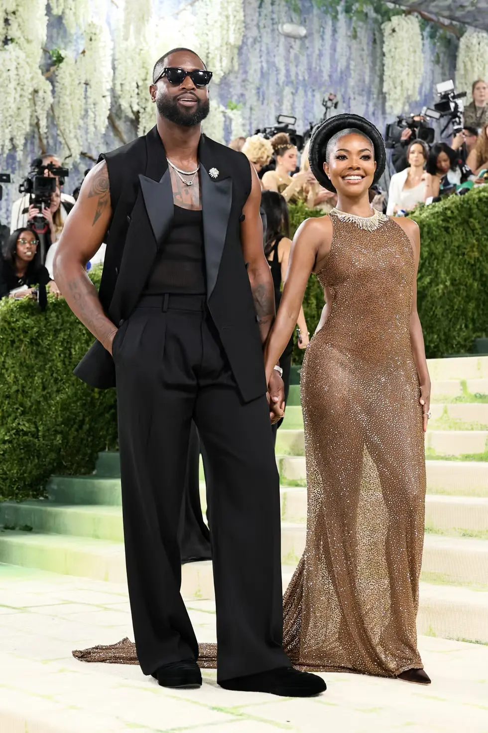 Dwyane wade - the game Couple on a green-carpeted stairway at a formal event; man in a black sleeveless vest and wide-legged pants with sunglasses, woman in a bronze sequined gown with a head wrap, holding hands.