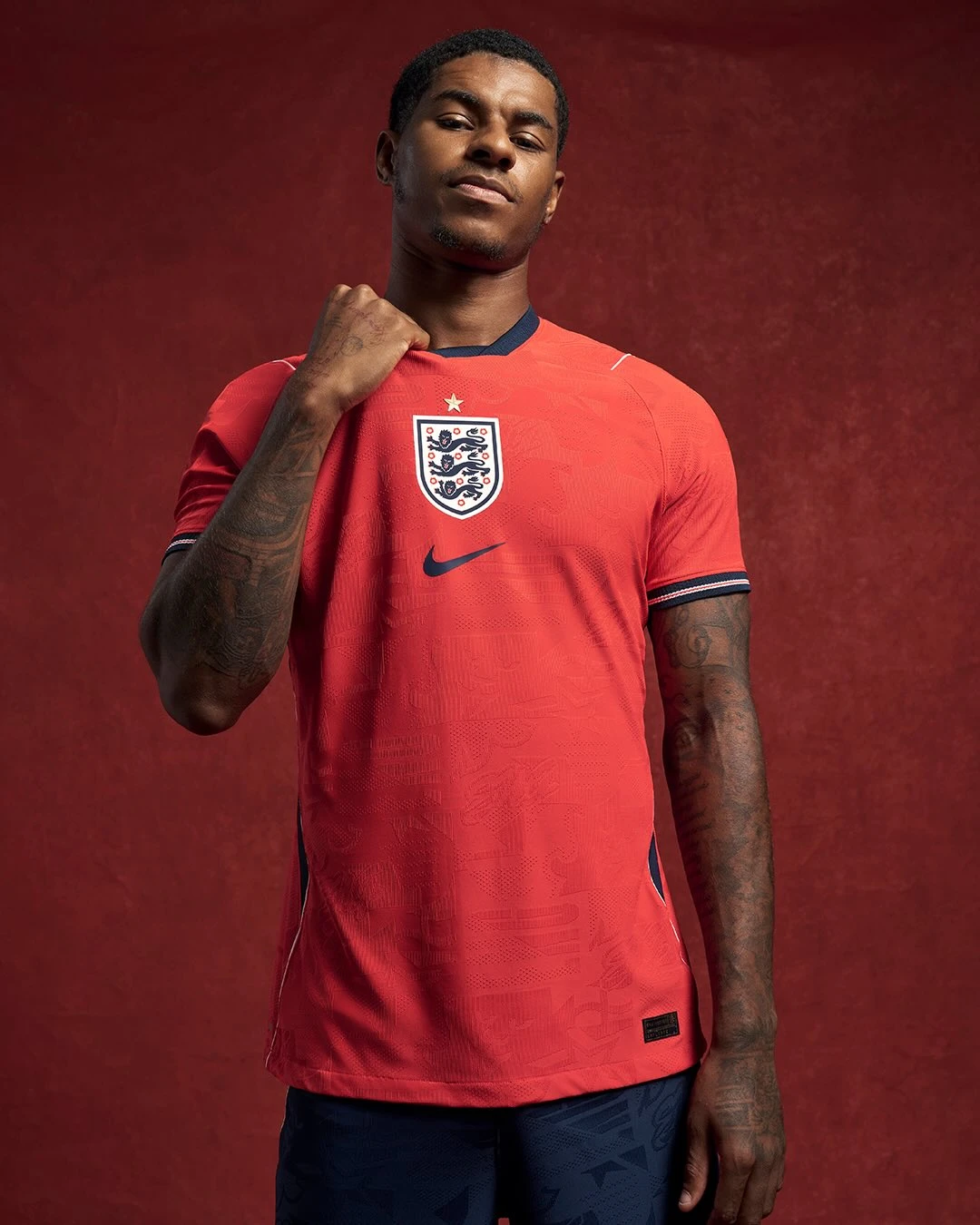 England away - the game Portrait of a young man in a red England soccer jersey, adjusting the collar against a red backdrop.