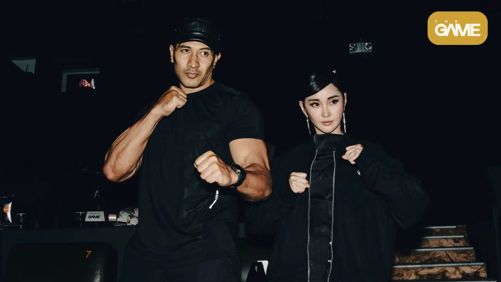 Eruption and alodia - the game Man and woman in black outfits strike a fighting pose in a dim venue, The Game logo in the top-right corner.