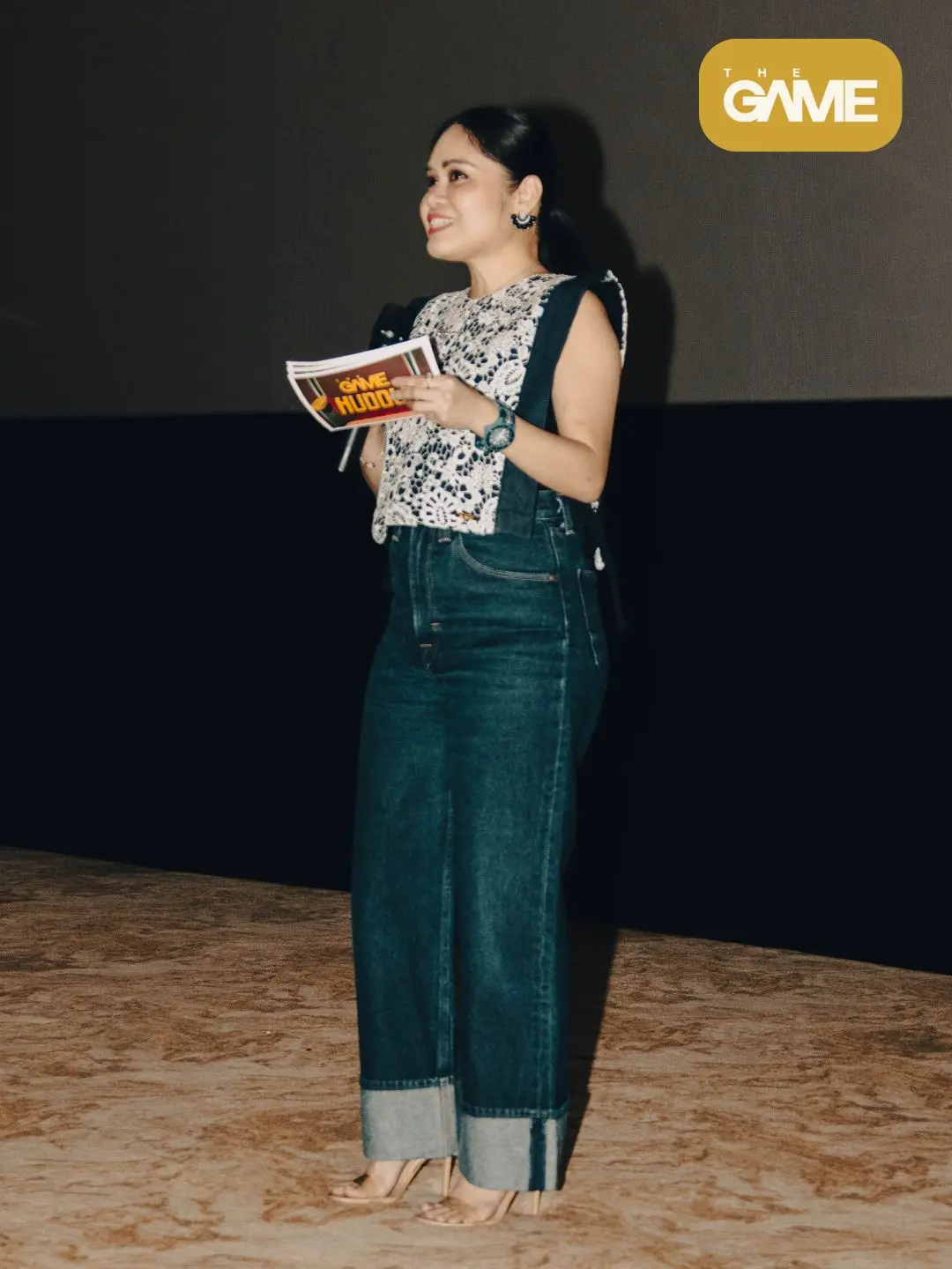 Ggb - the game Woman presenter on stage holding a booklet, wearing a white lace top and dark wide‑leg jeans with heeled sandals; The Game logo appears in the top-right corner of the image.