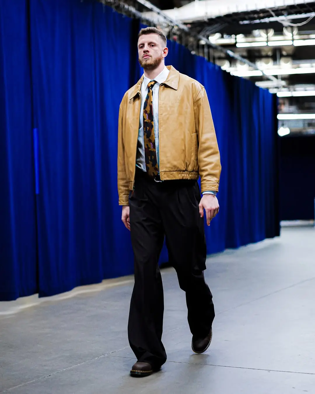 Isaiah hartenstein - the game Male model walking down a hallway with blue curtains, wearing a tan bomber jacket, white shirt, camo tie, and black trousers.